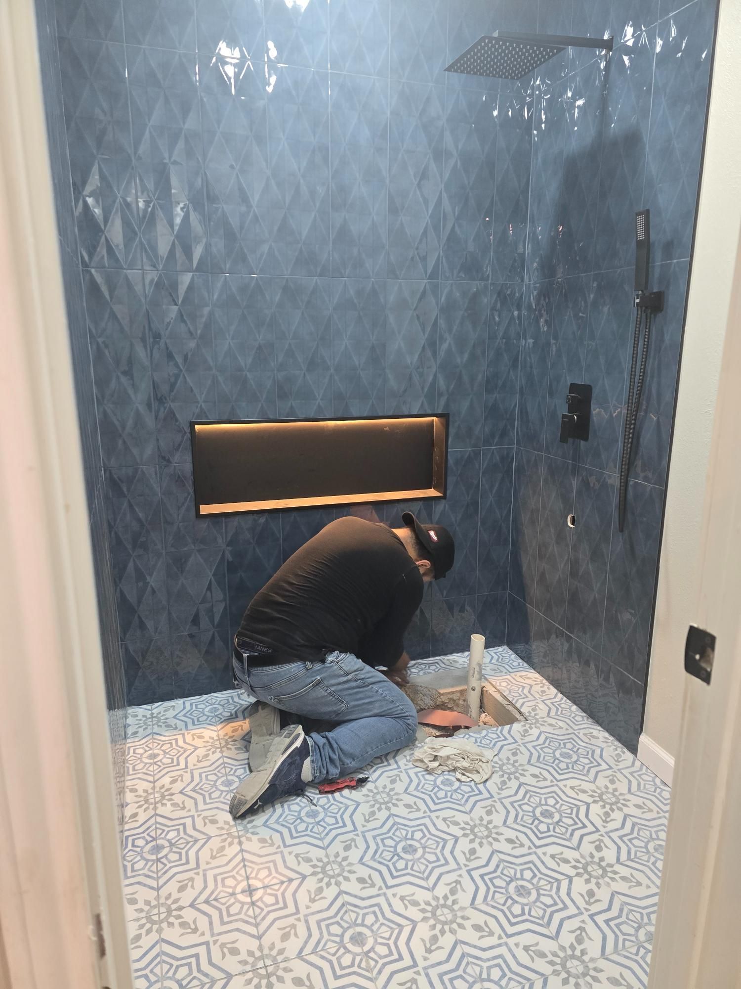 Person tiling a blue patterned shower floor. Blue diamond-patterned tile on walls; recessed shelf.
