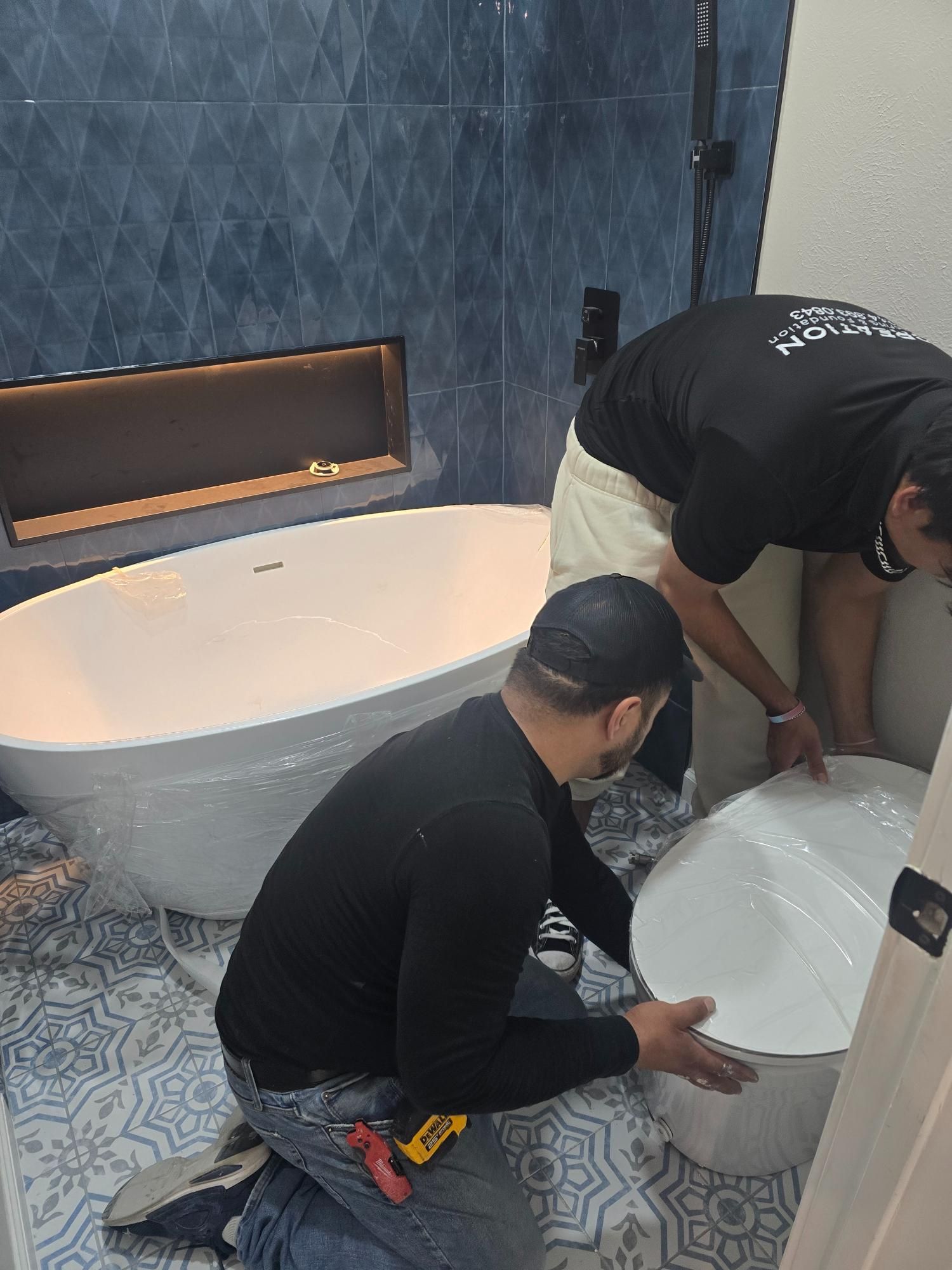 Two people installing a toilet in a newly renovated bathroom with a bathtub, blue tile, and patterned floor.