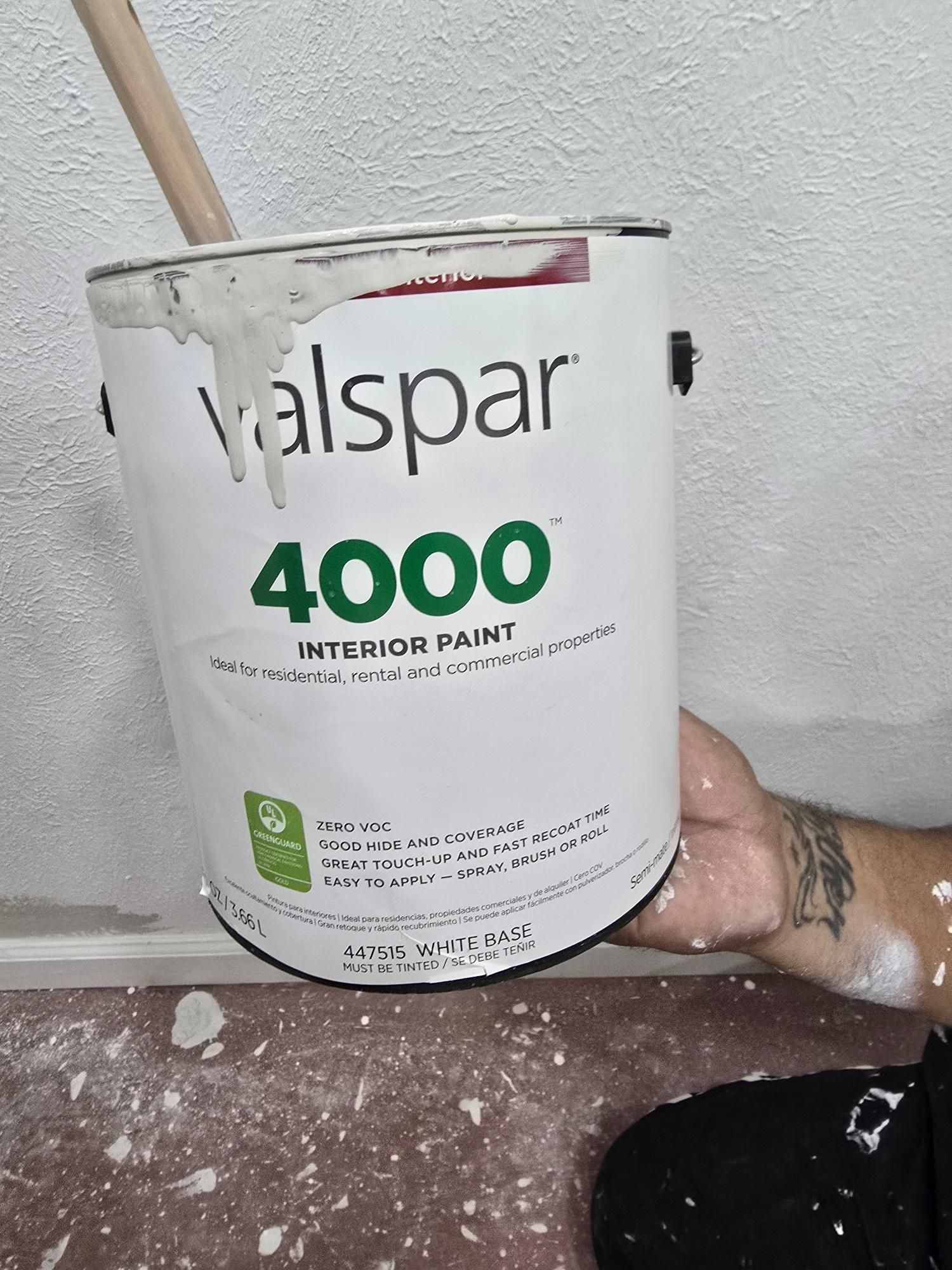 Hand holding a Valspar 4000 interior paint can with a wooden stirrer. Paint drips down the side, on a white wall.