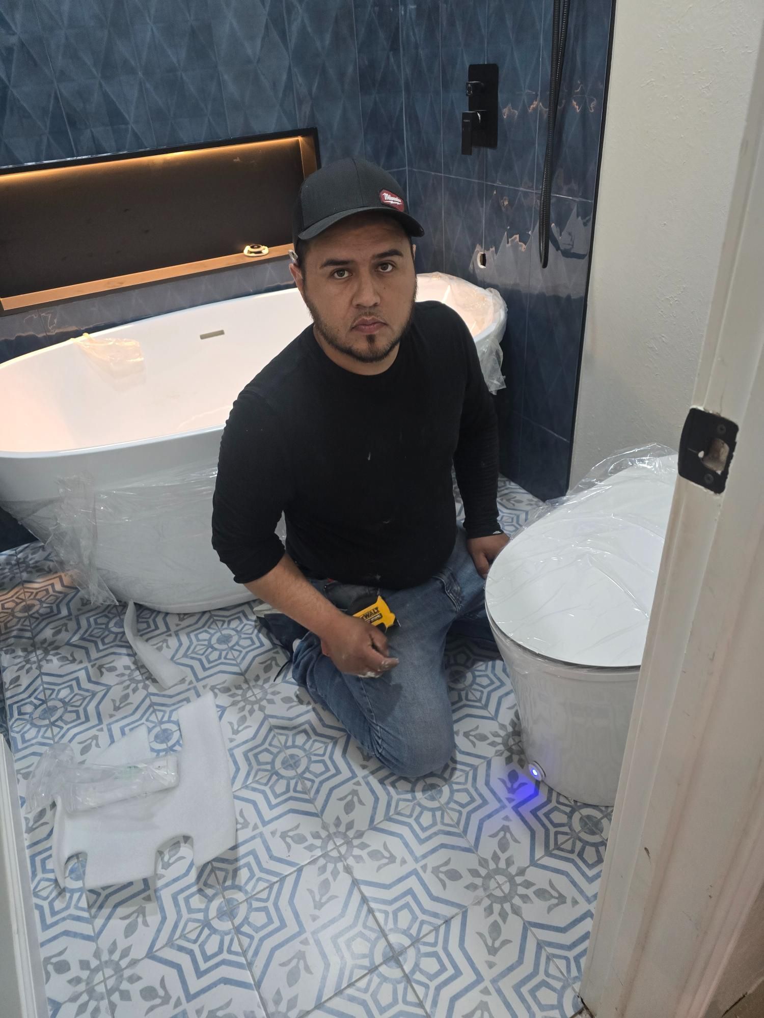 Man kneeling in bathroom, installing toilet. Blue patterned floor and walls, white tub.