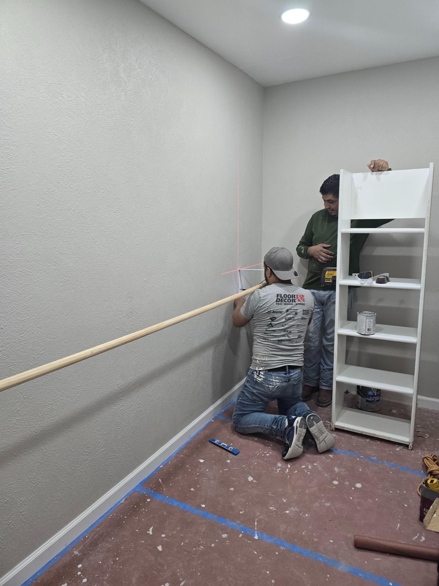 Two people installing a wooden rod on a textured gray wall inside a room.