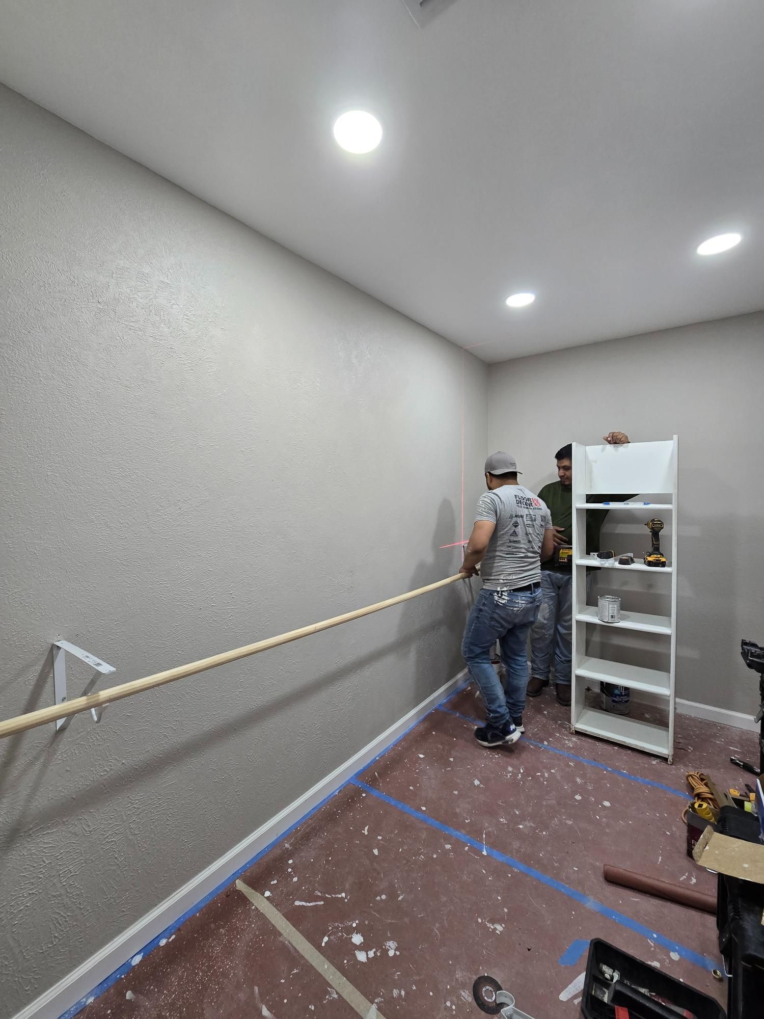 Two people installing a wooden handrail on a textured gray wall, inside a room under construction.