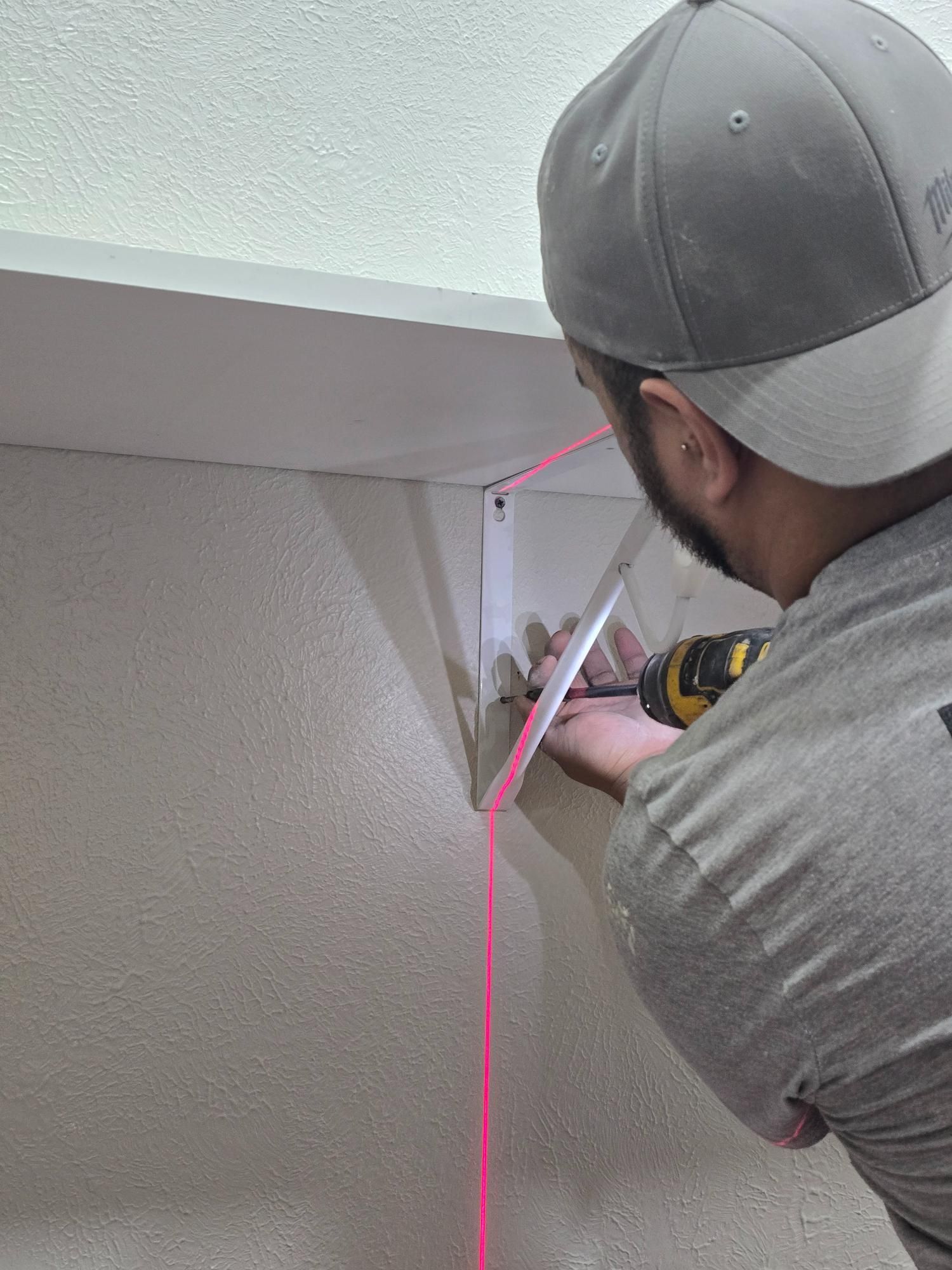 A person uses a drill to install a corner bracket; a pink laser line guides the work.