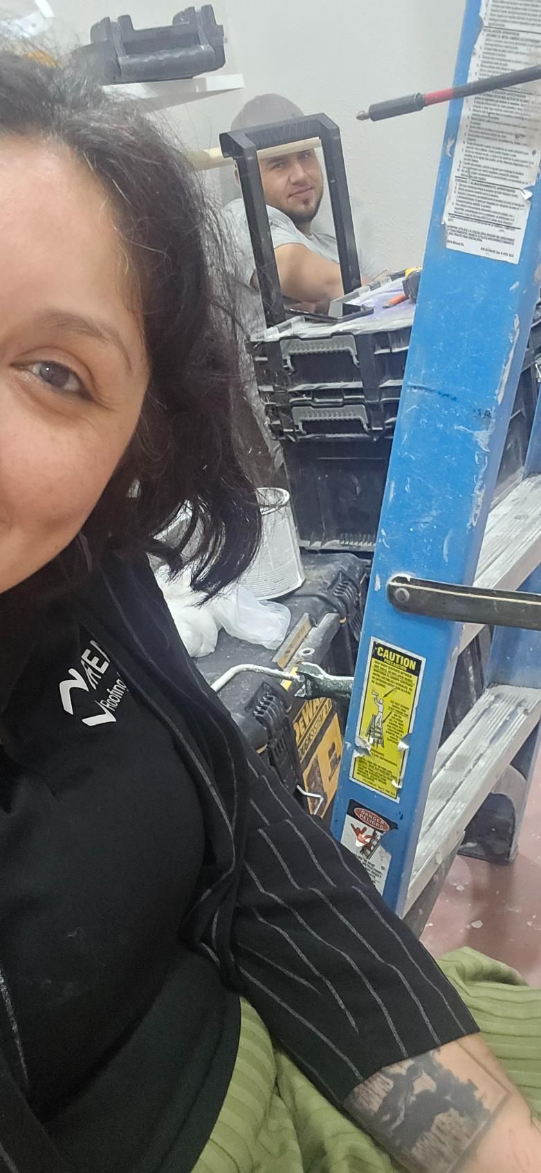 Woman taking a selfie; another person in background. Blue ladder, gray hardhat, black jacket, and tattoos are visible.