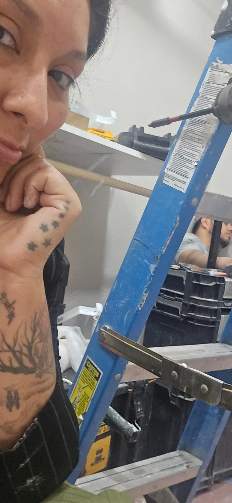 Person next to a blue ladder in a workshop; a clamp is attached to the ladder. The person has tattoos on their arm.
