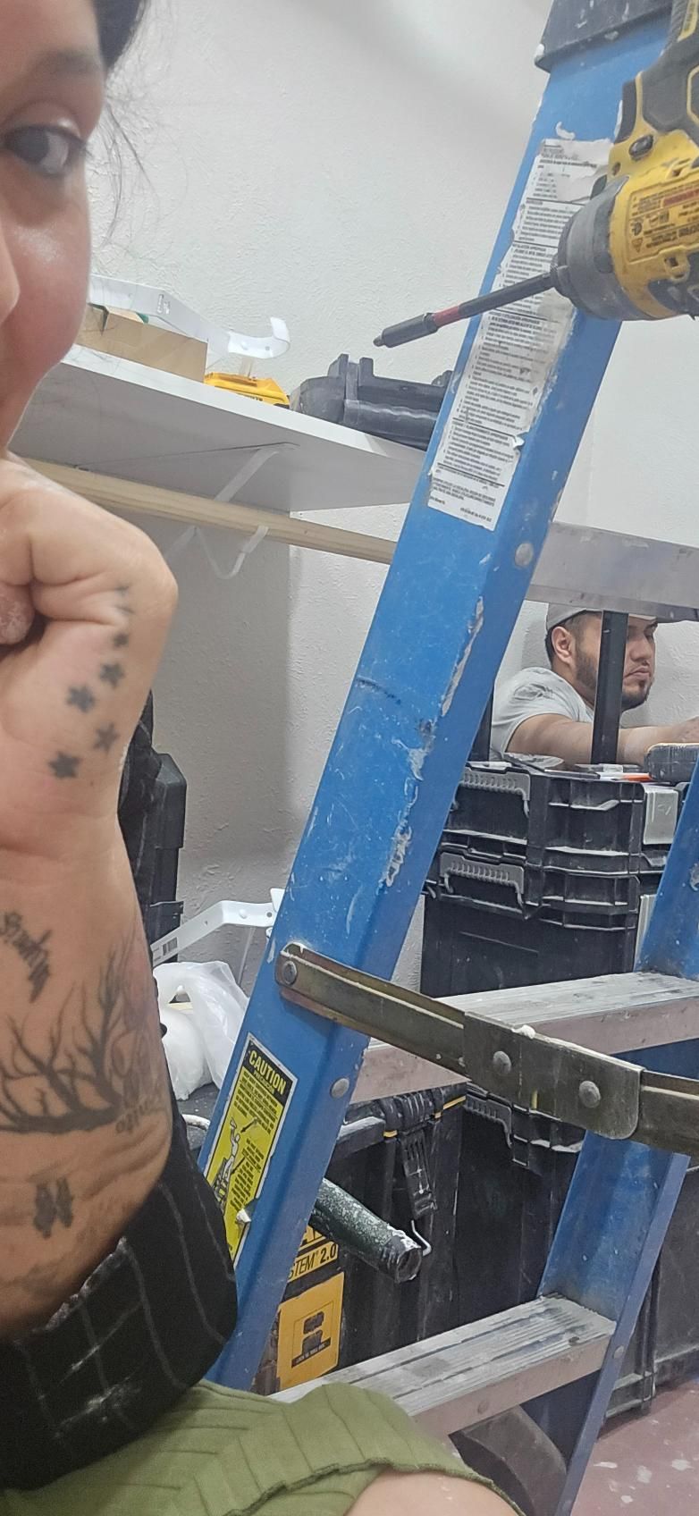 Person with arm tattoos smiles at the camera, standing near a blue ladder in a room with a worker in the background.