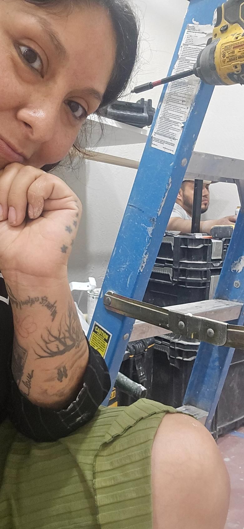 Person with arm tattoos poses near blue ladder; another person in background.