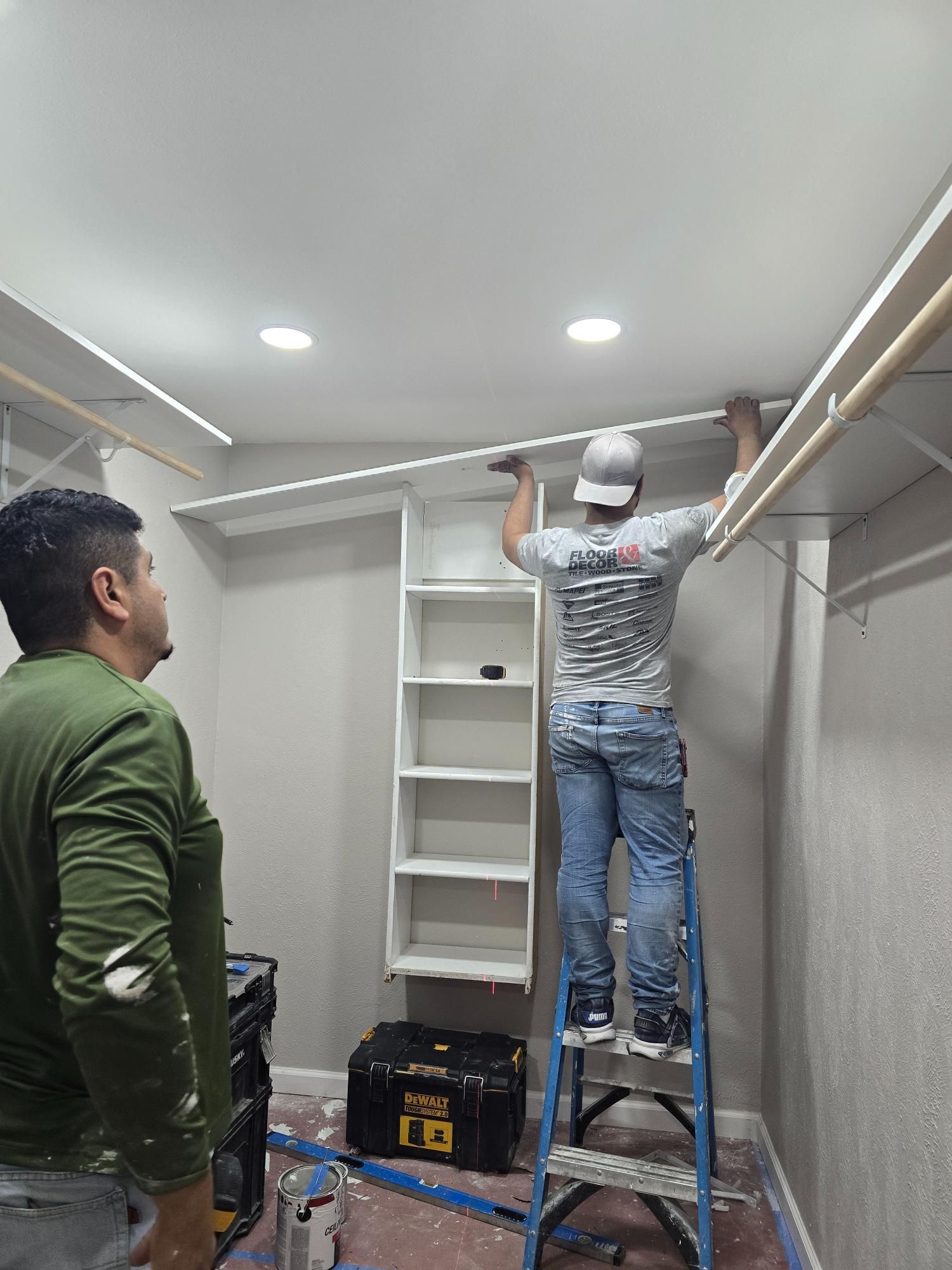 Two men installing a closet shelf. One stands on a ladder, the other observes. The closet is white with a gray textured wall.