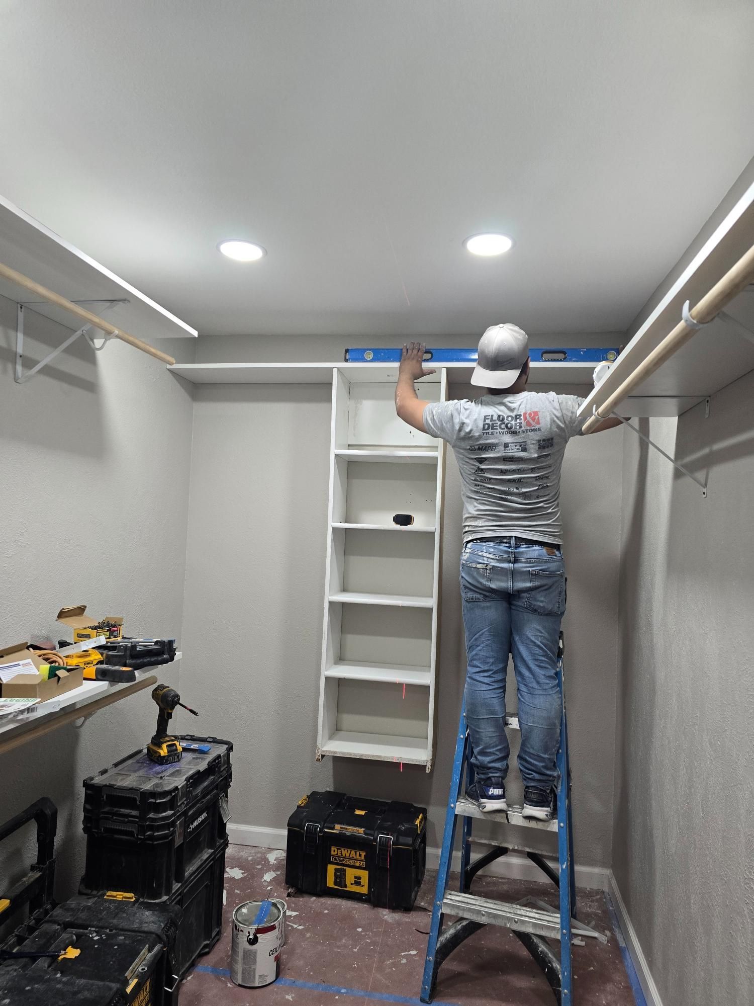 Person on ladder installing closet shelves in a room with tools on the floor.