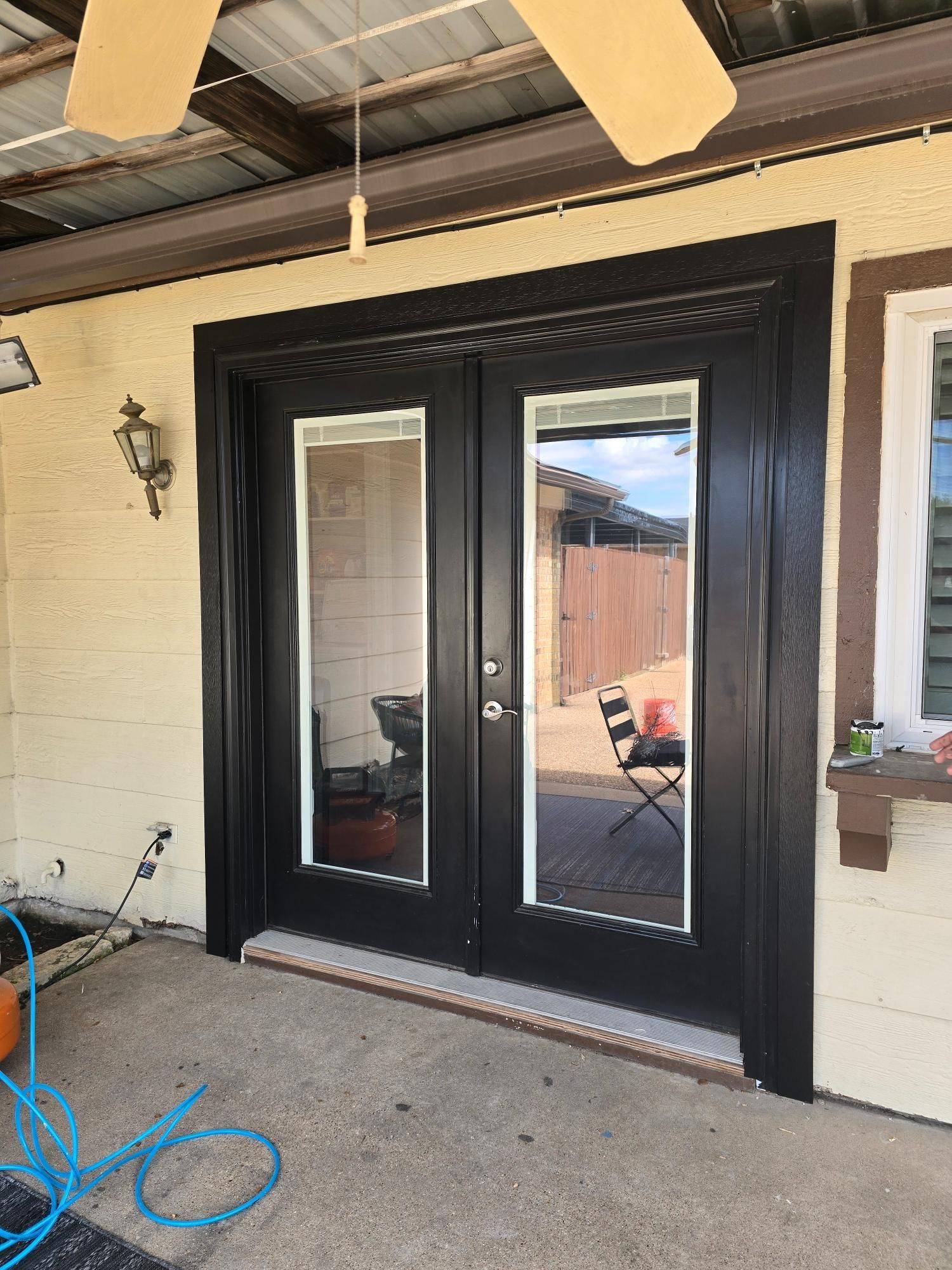 Black double doors with glass panels and a dark brown frame, set in an outdoor space with tan walls.