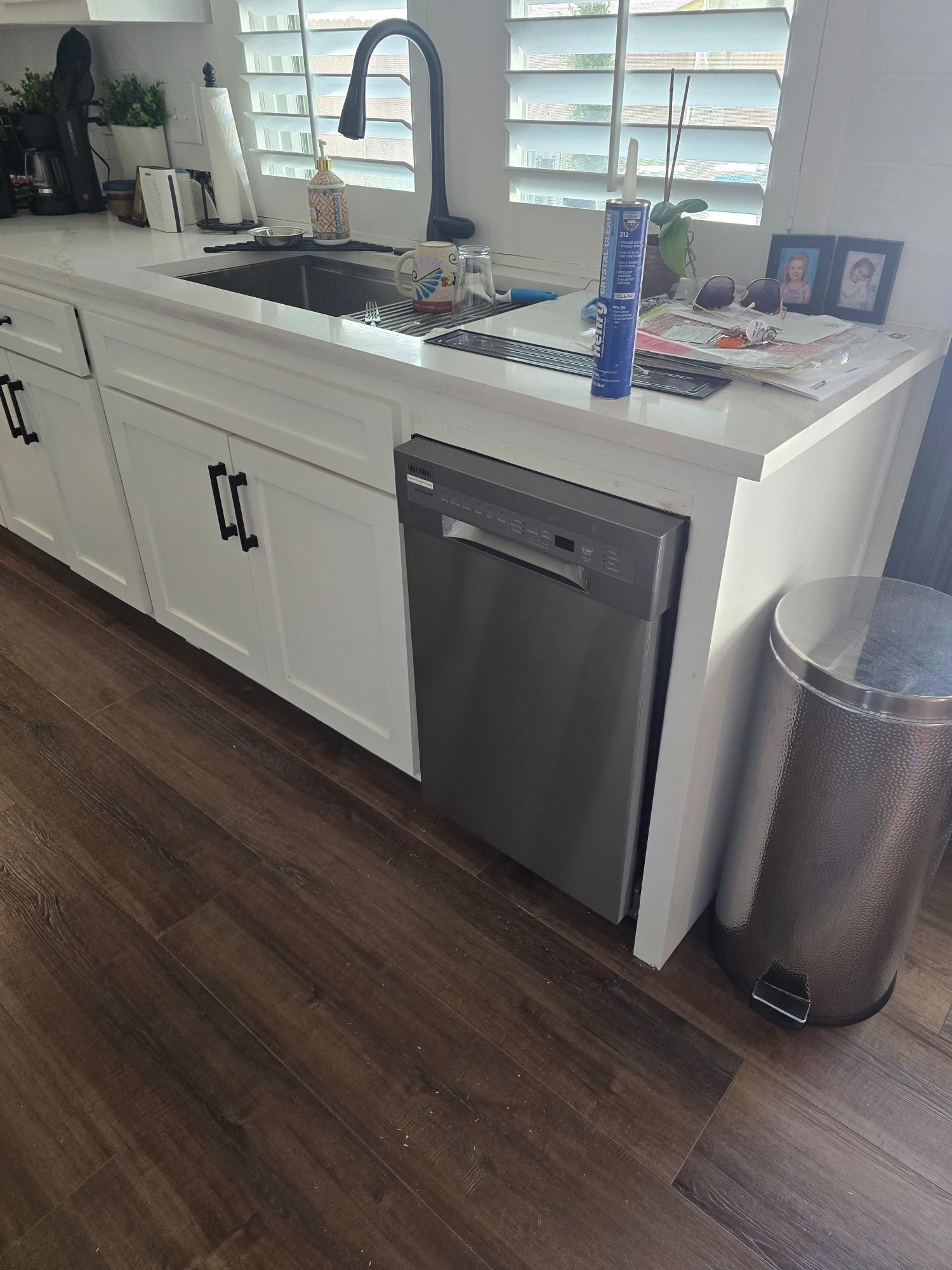 Kitchen counter with sink and dishwasher. Silver trash can sits to the right. Dark wood floor.