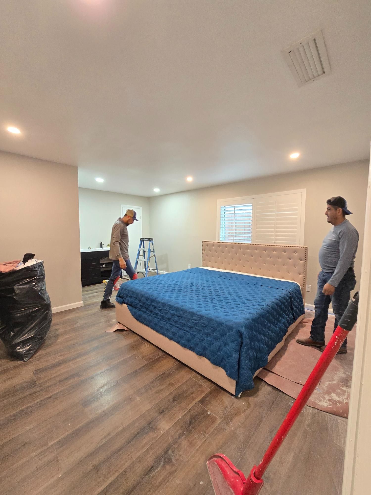 Two people move a bed in a room with wood floors and a dark cabinet. A trash bag and a broom are nearby.