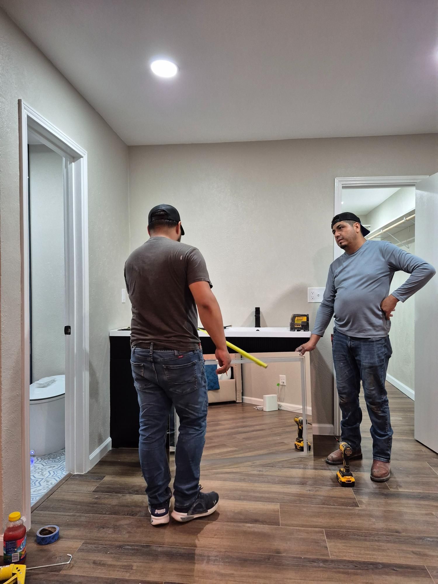 Two men in a room, one holding a measuring tape, the other pointing, with tools and a doorway visible.
