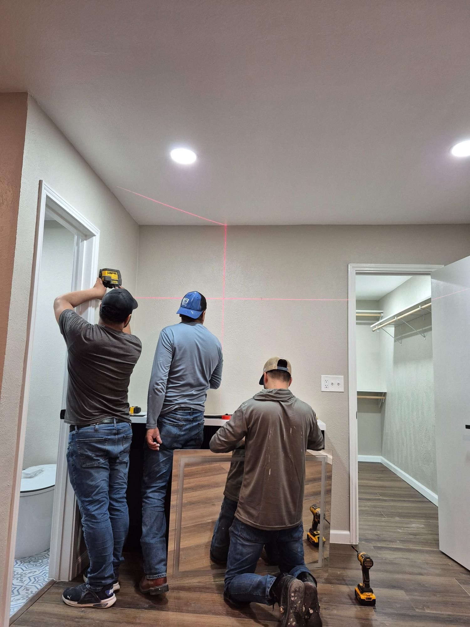 Three people installing cabinetry using a laser level in a room with hardwood floors.