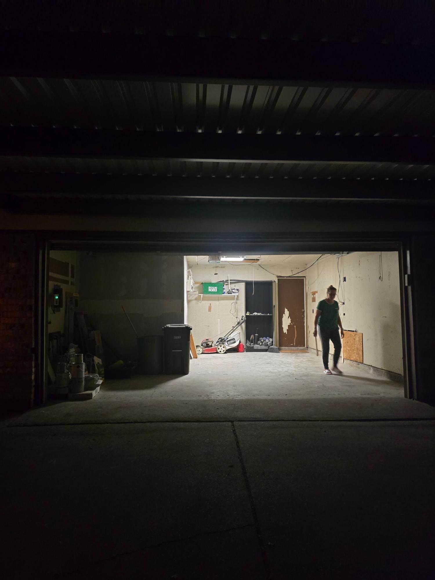A person stands in a dimly lit garage. The garage door is open, revealing items inside.