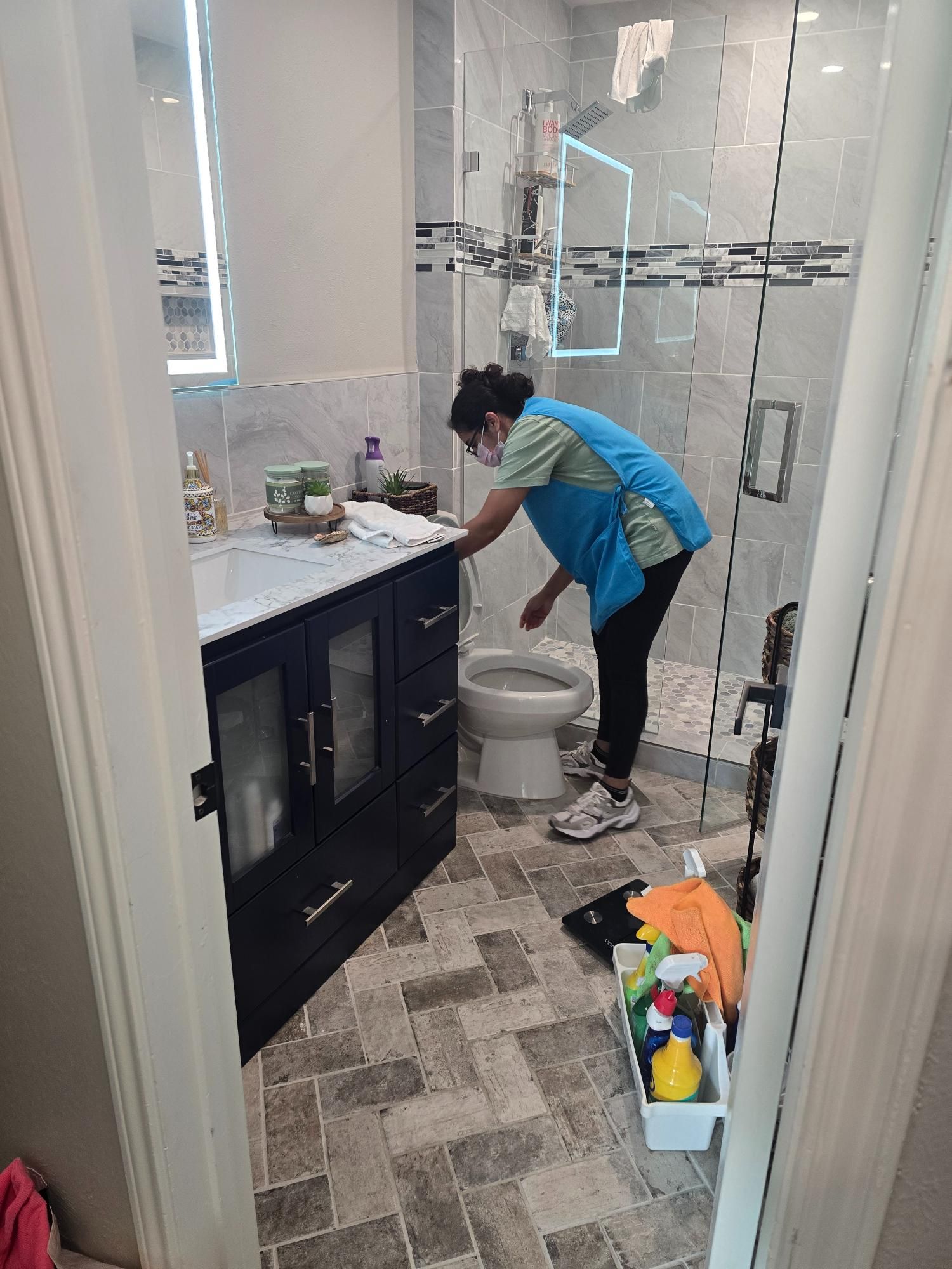 Person cleaning a modern bathroom. They wear a mask and blue apron, cleaning near the toilet.