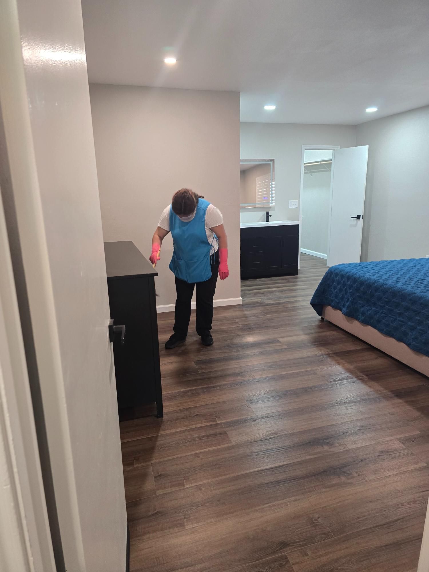Person cleaning a bedroom with dark wood floors, wearing a blue apron and pink gloves.