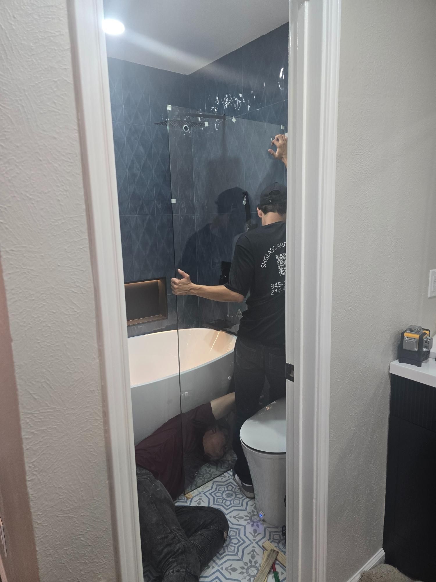 People installing glass shower door in a bathroom with a white tub and patterned floor.