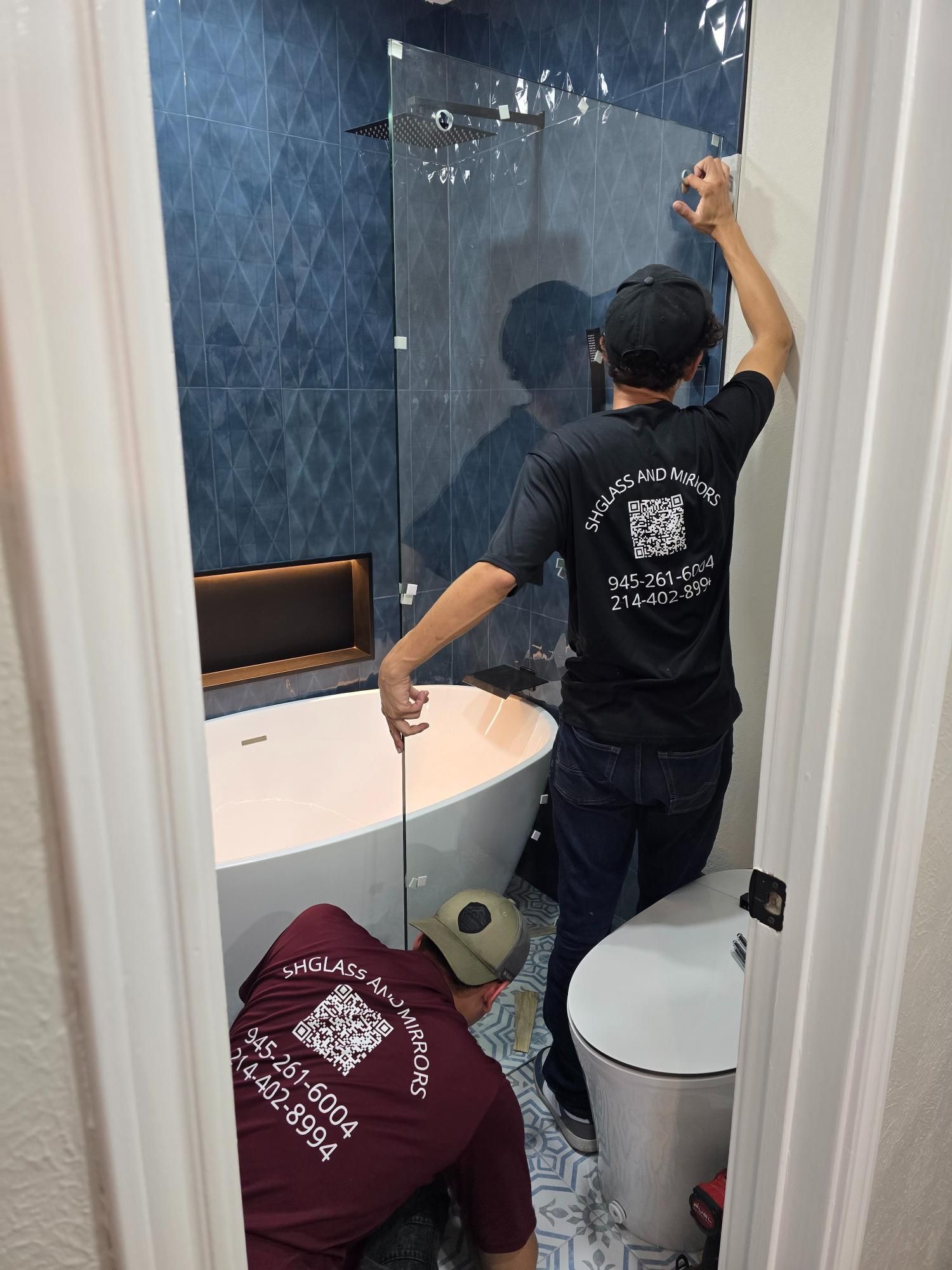Two workers installing a glass shower door in a blue-tiled bathroom with a bathtub and toilet.