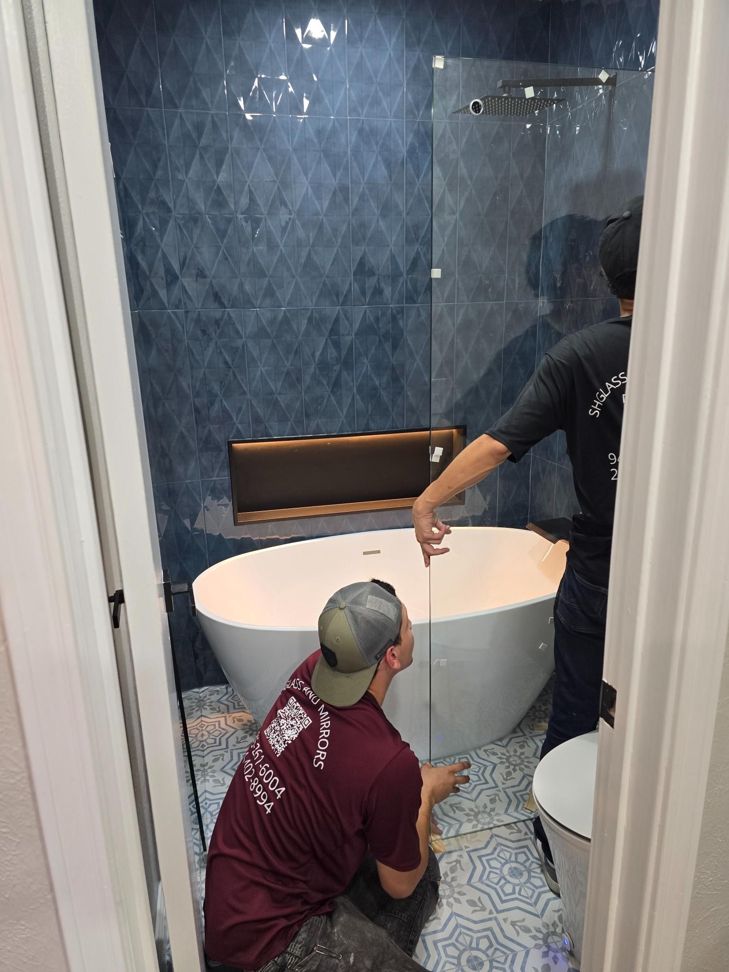 Two people installing a shower glass panel in a bathroom with blue tile and a white tub.