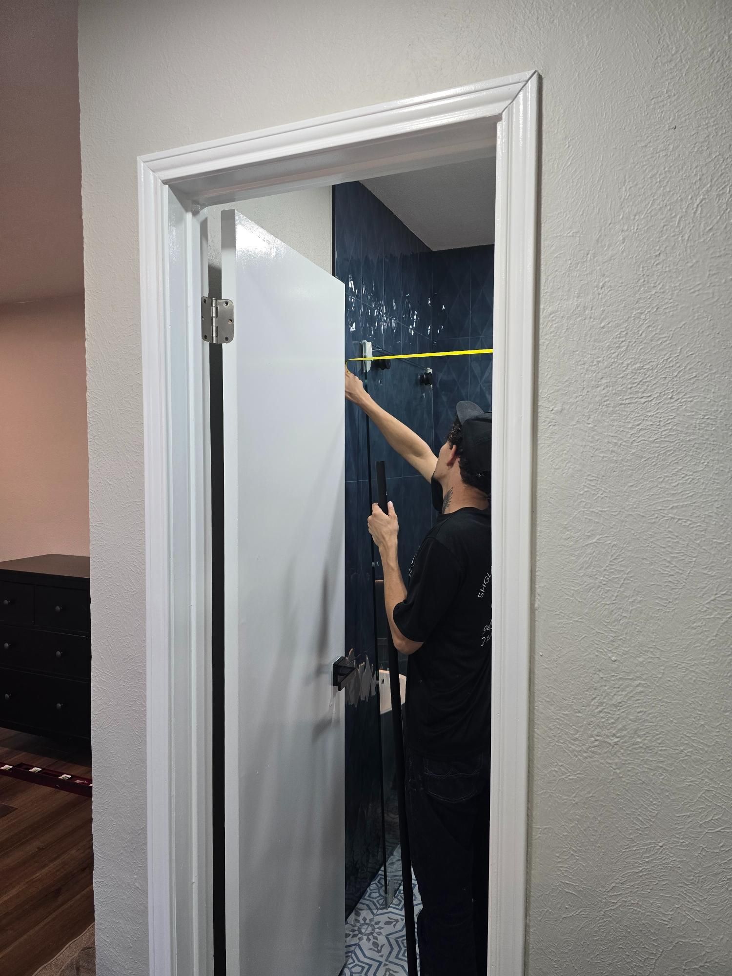 Man cleaning a shower with a sprayer inside a white-framed doorway; blue tile visible.