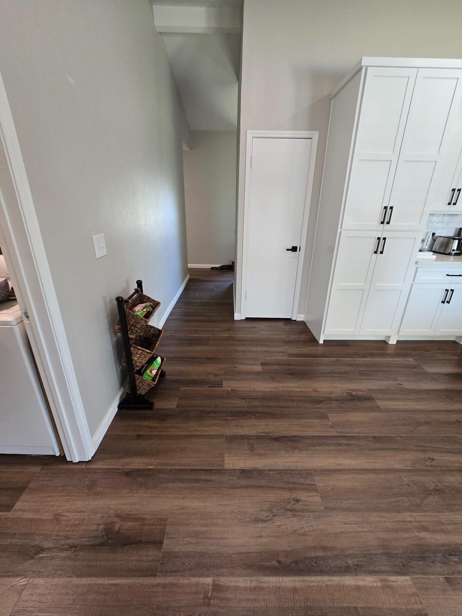 Kitchen with dark wood flooring, tall white cabinets, a small produce rack, and a closed white door.