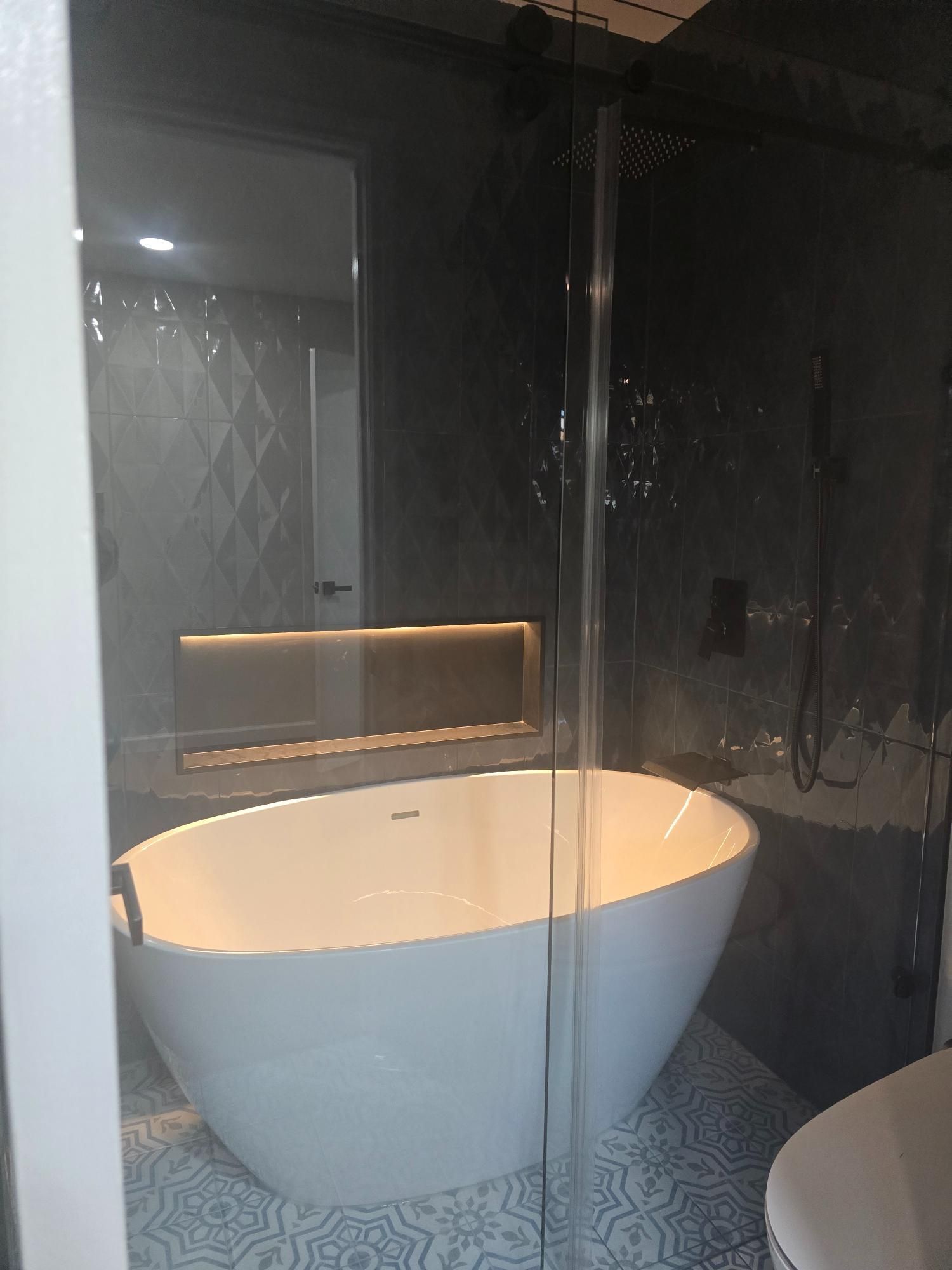 Modern bathroom with white oval tub, glass shower, patterned tile floor, and dark gray walls.