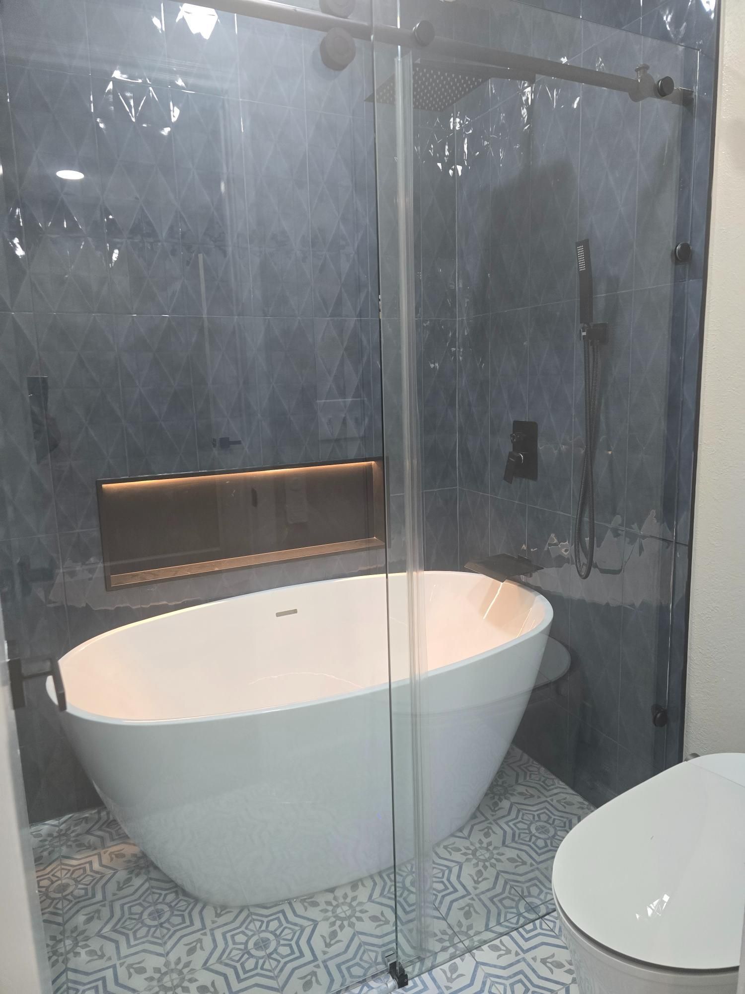 Modern bathroom with white tub, glass shower, patterned tile floor, and dark blue wall tiles.