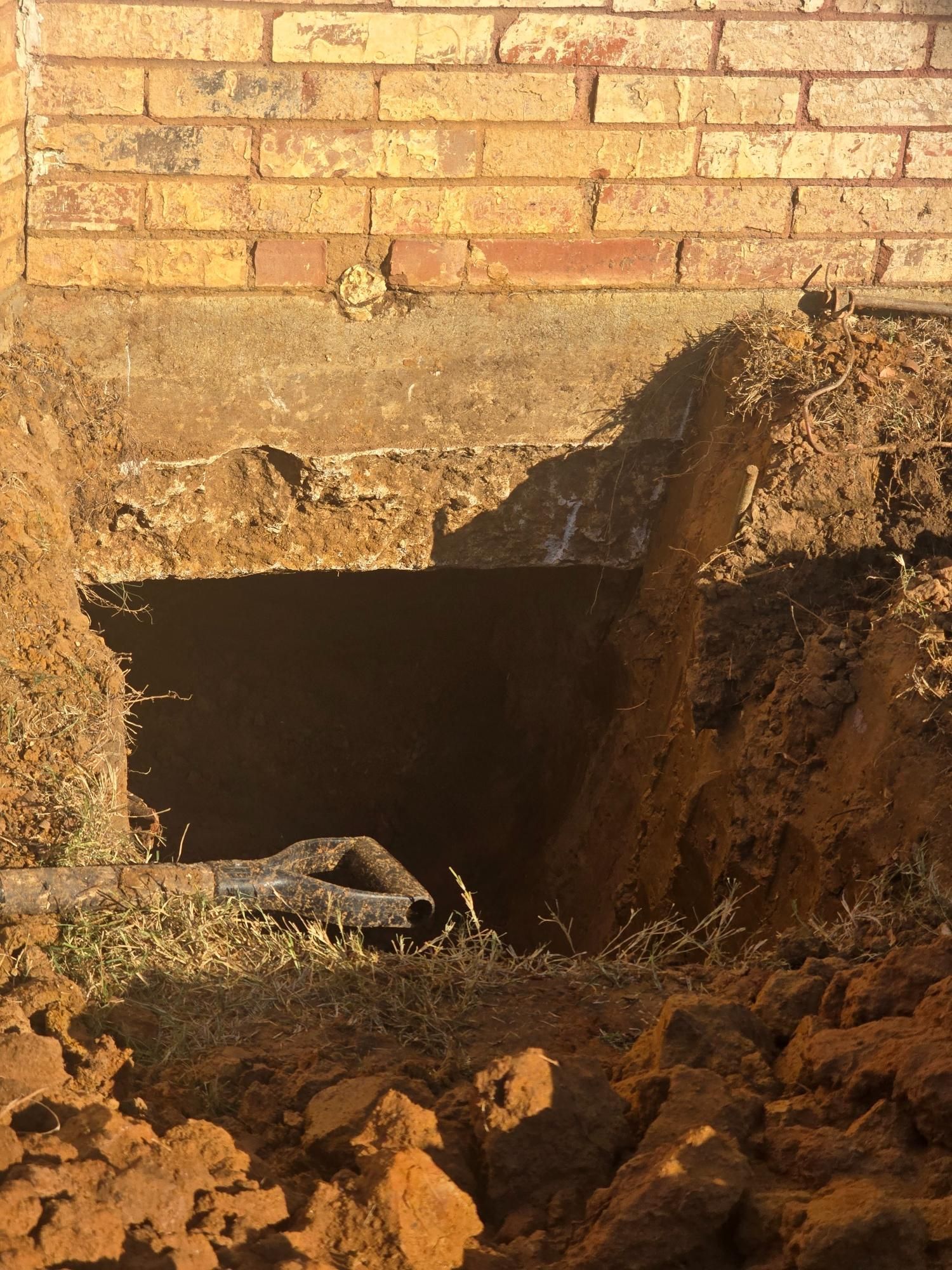 Hole dug near brick foundation, revealing a dark crawl space. Brown soil, orange brick, and a pipe visible.