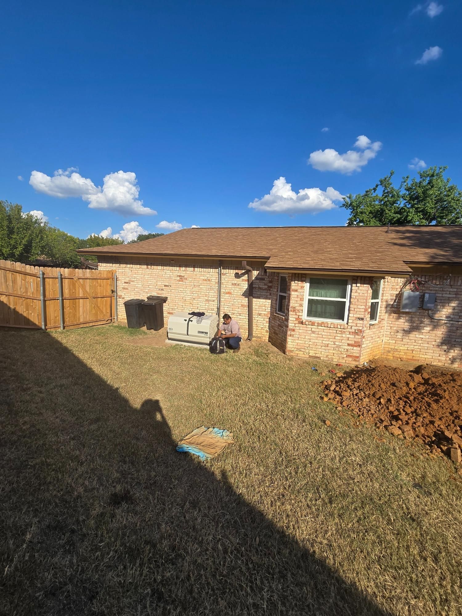 Backyard view of a house, wooden fence, brown grass, blue sky with clouds. A person sits near the brick wall.