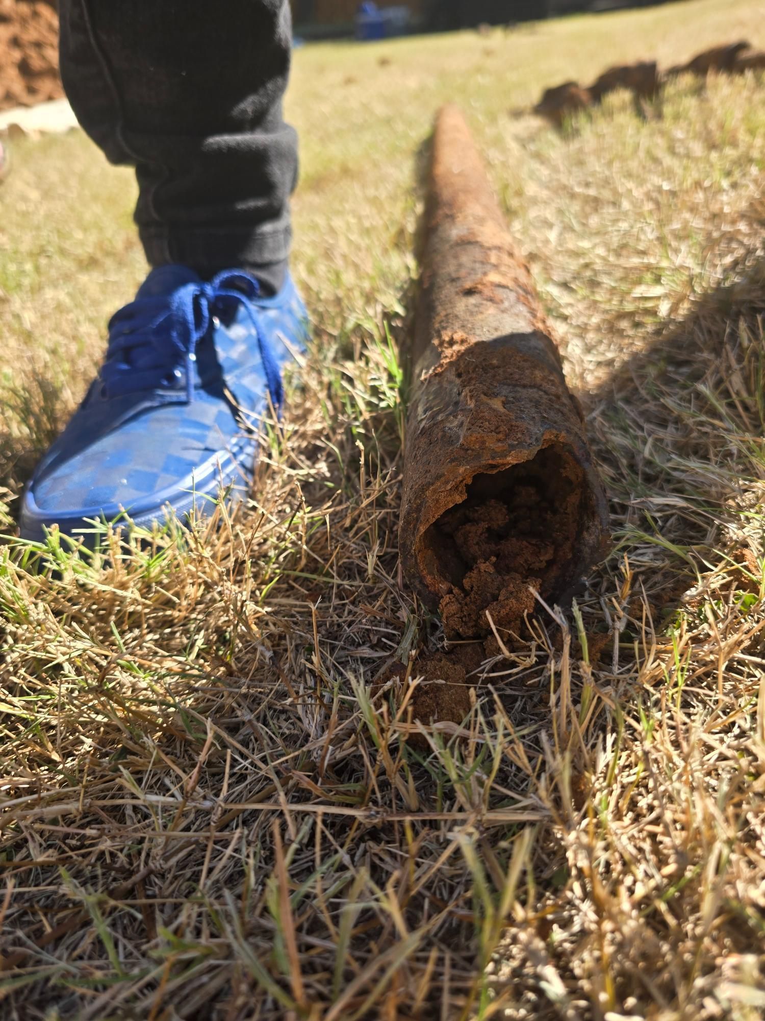 A rusty pipe lying on grass with a person's blue shoe nearby.