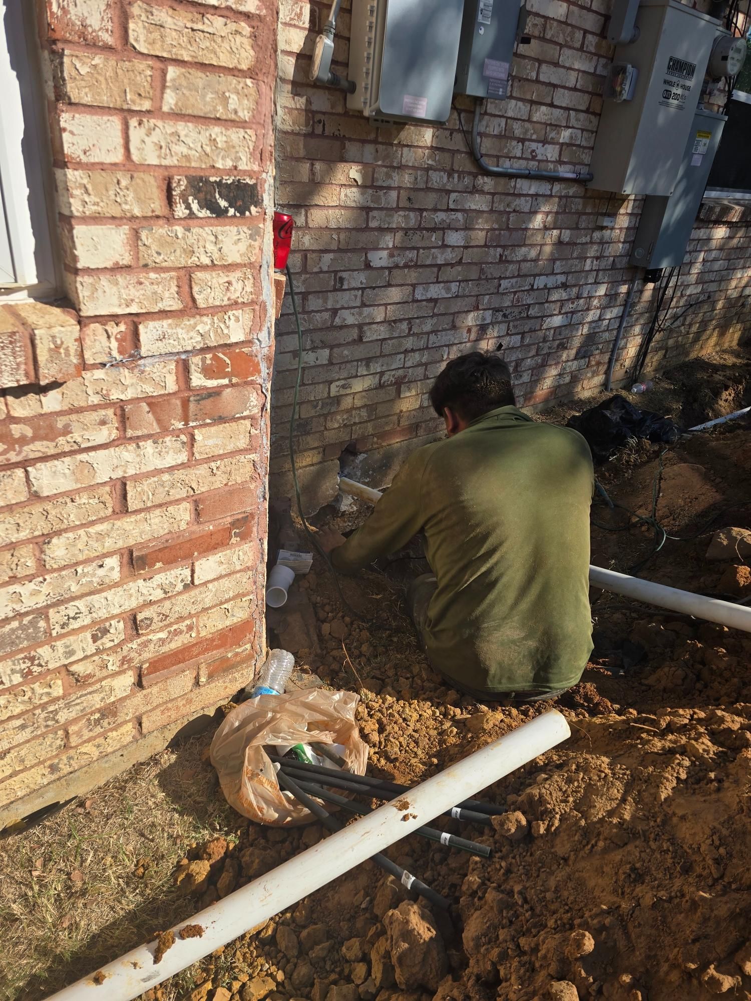 Person kneeling near a brick building, working on underground pipes in a trench.