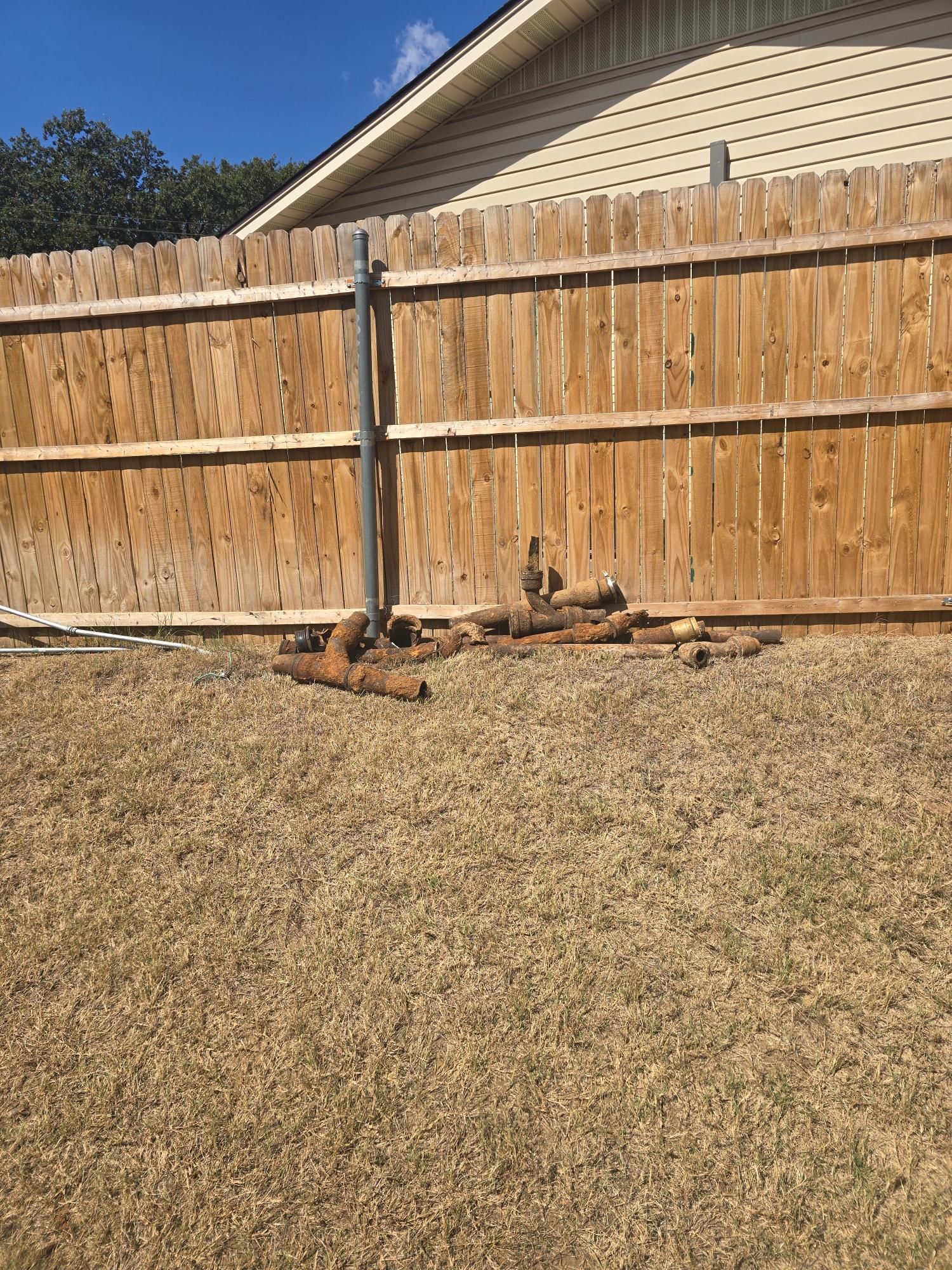 A disassembled metal pole structure and various pipe pieces sit on brown grass against a wooden fence.