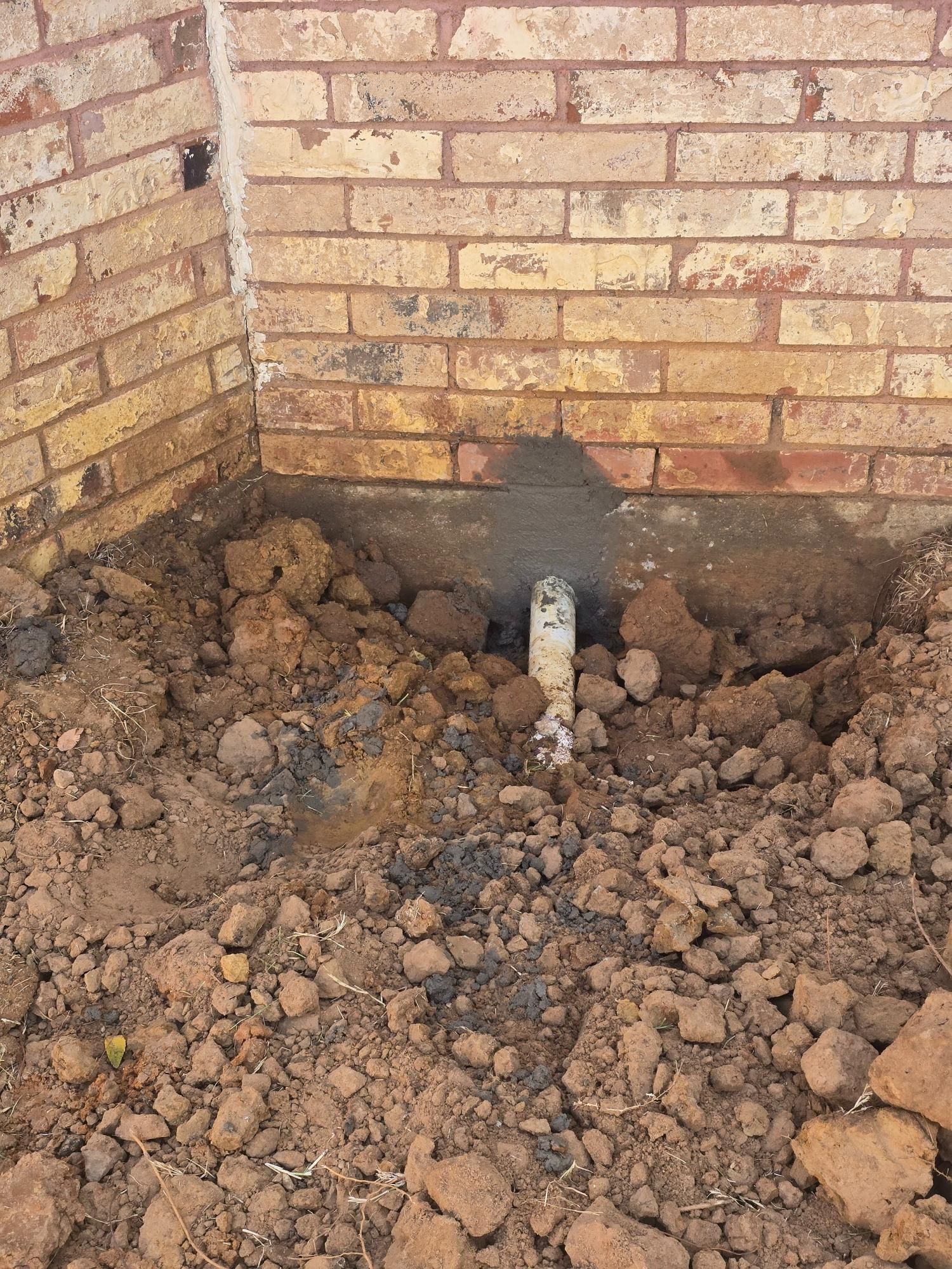 A white pipe emerging from the dirt near a brick wall foundation.
