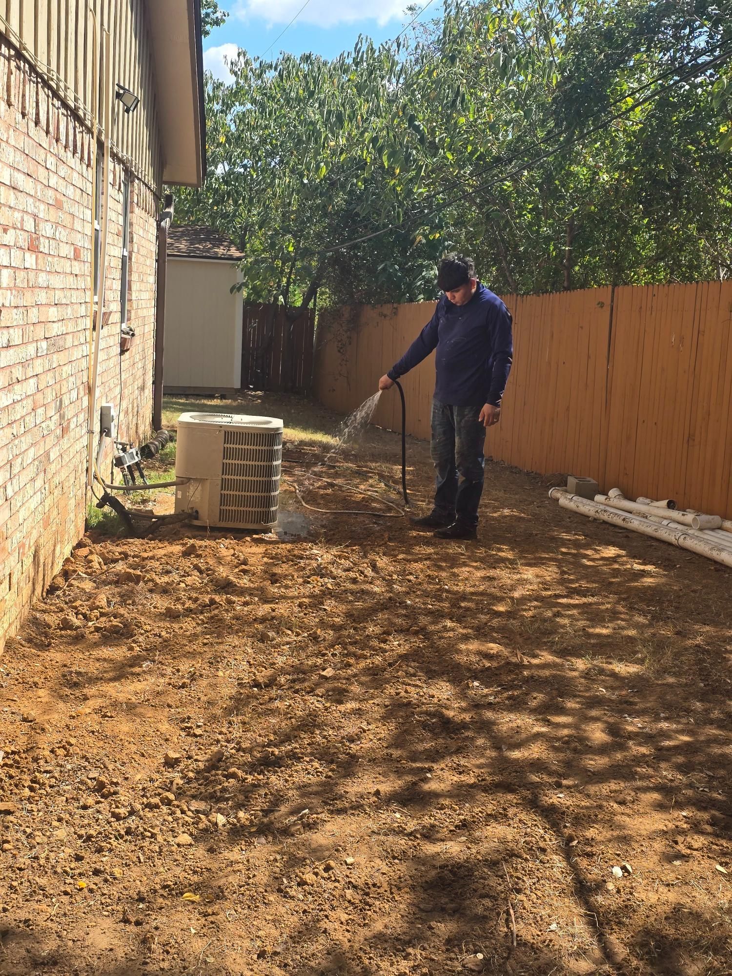 Person watering an exposed dirt area next to a building and fence on a sunny day.