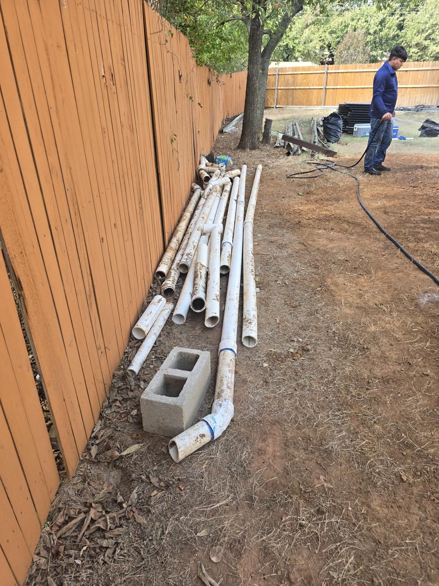 Pipes, a cinder block, and a person stand near a wooden fence on a brown grassy area.