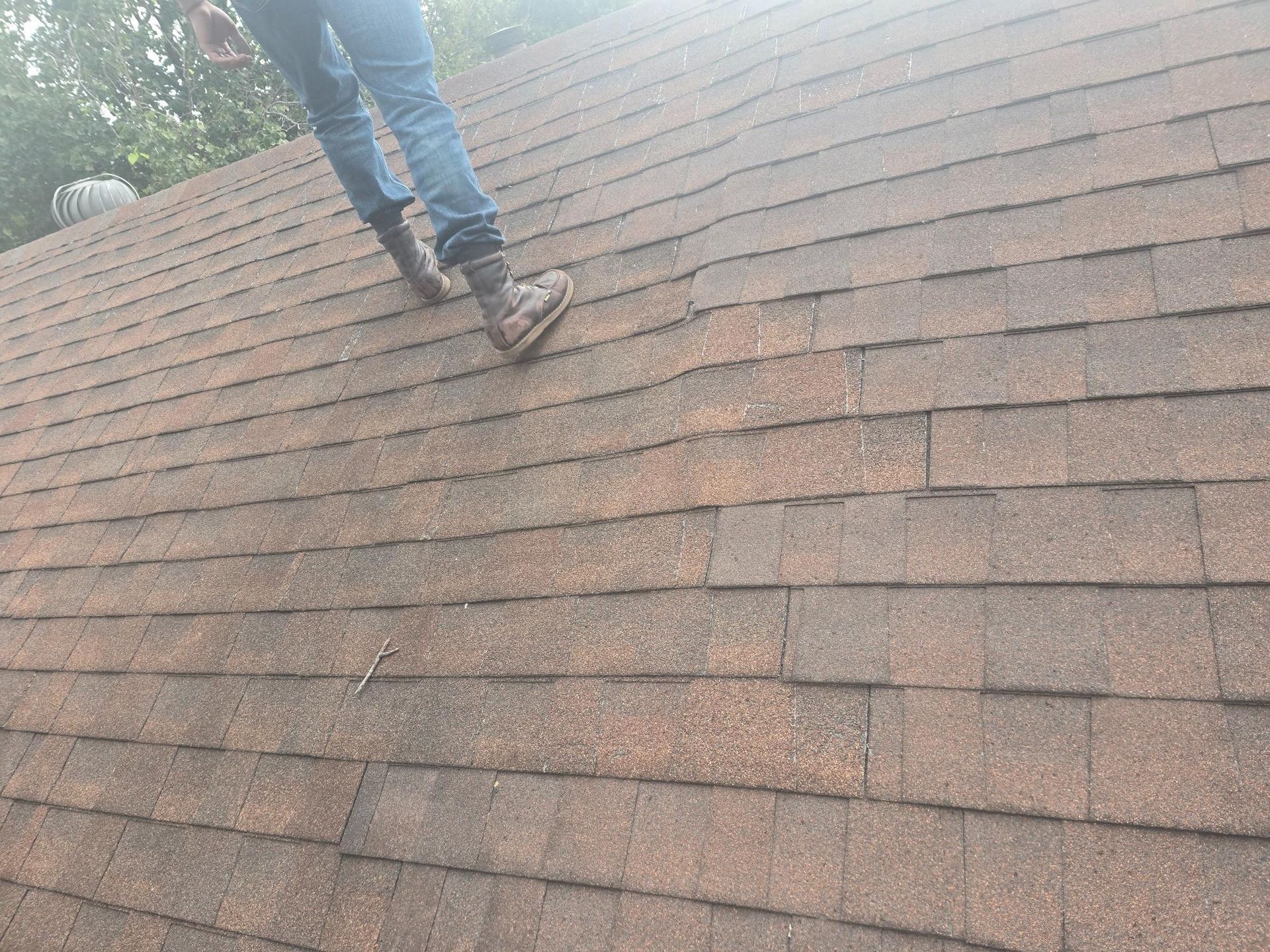 Person in jeans and work boots standing on a brown shingle roof.