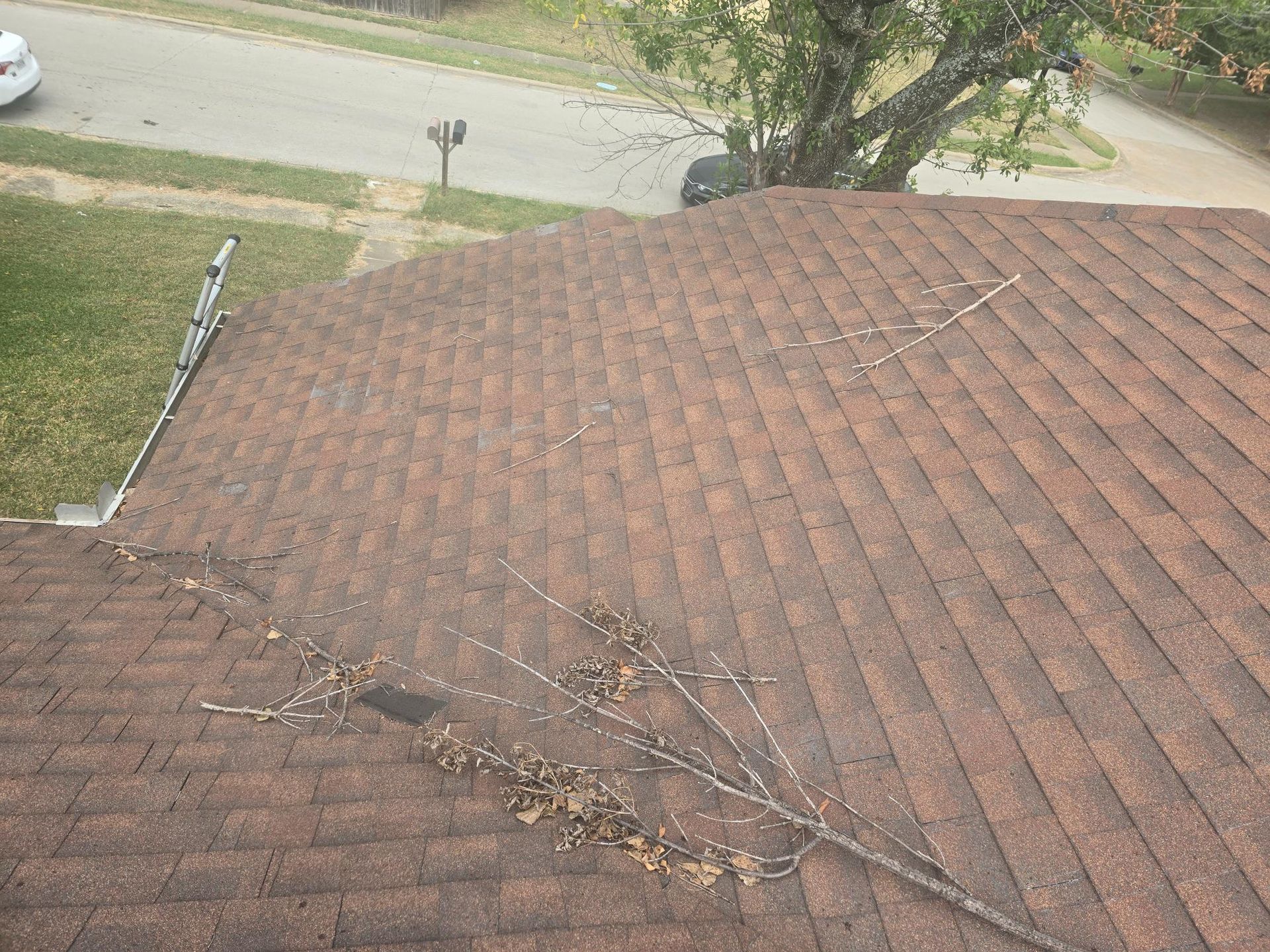 Brown asphalt shingle roof with a fallen tree branch. View from above with a street in the background.