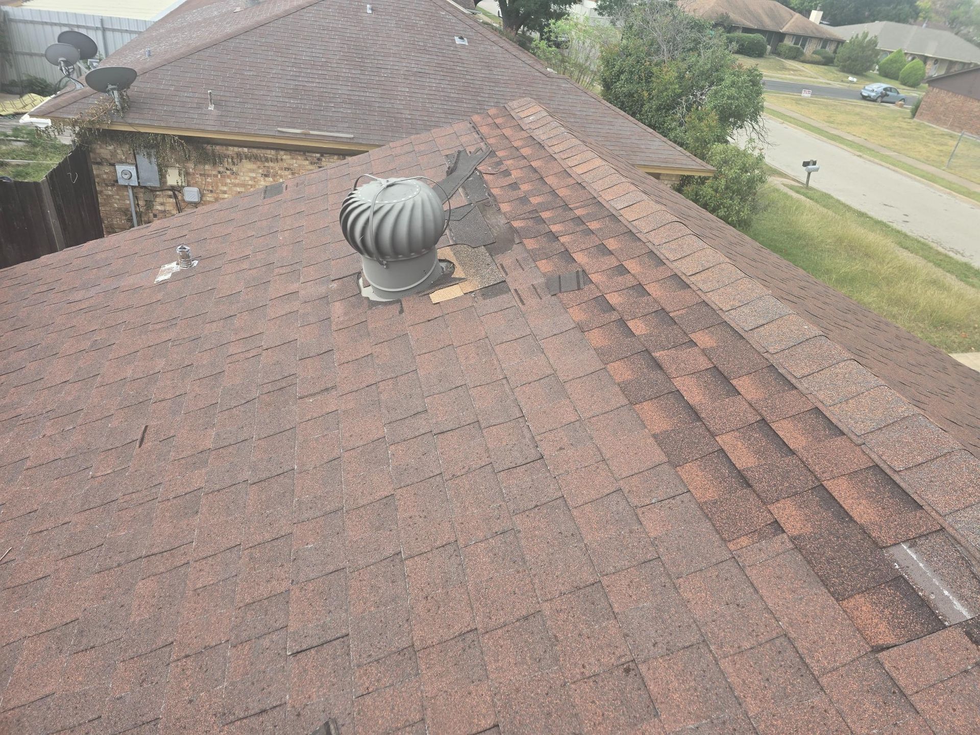Brown asphalt shingle roof with a vent and damaged shingles, next to green grass and a street.