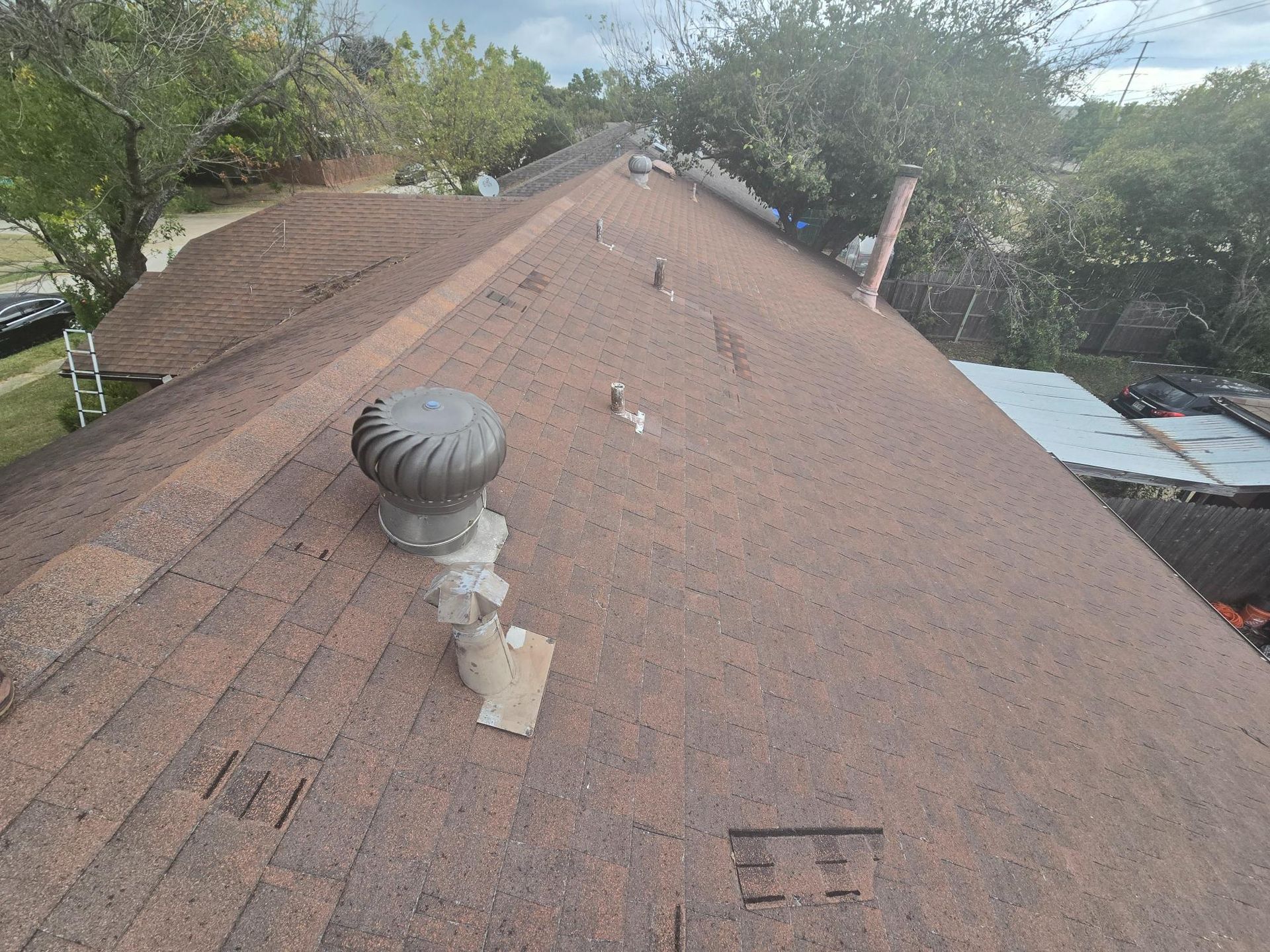 Brown asphalt shingle roof with a vent and damaged shingles, trees in the background, daytime.