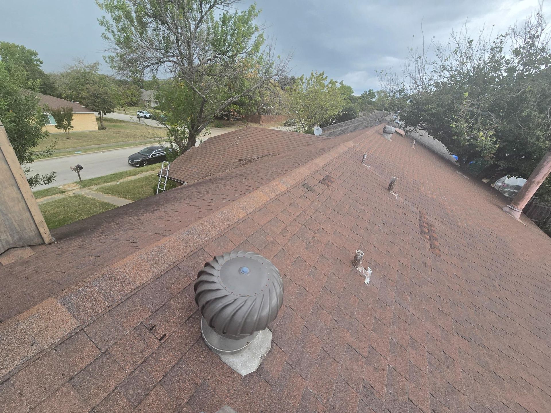 View from rooftop with brown shingles, a turbine vent, and houses in the distance under a cloudy sky.