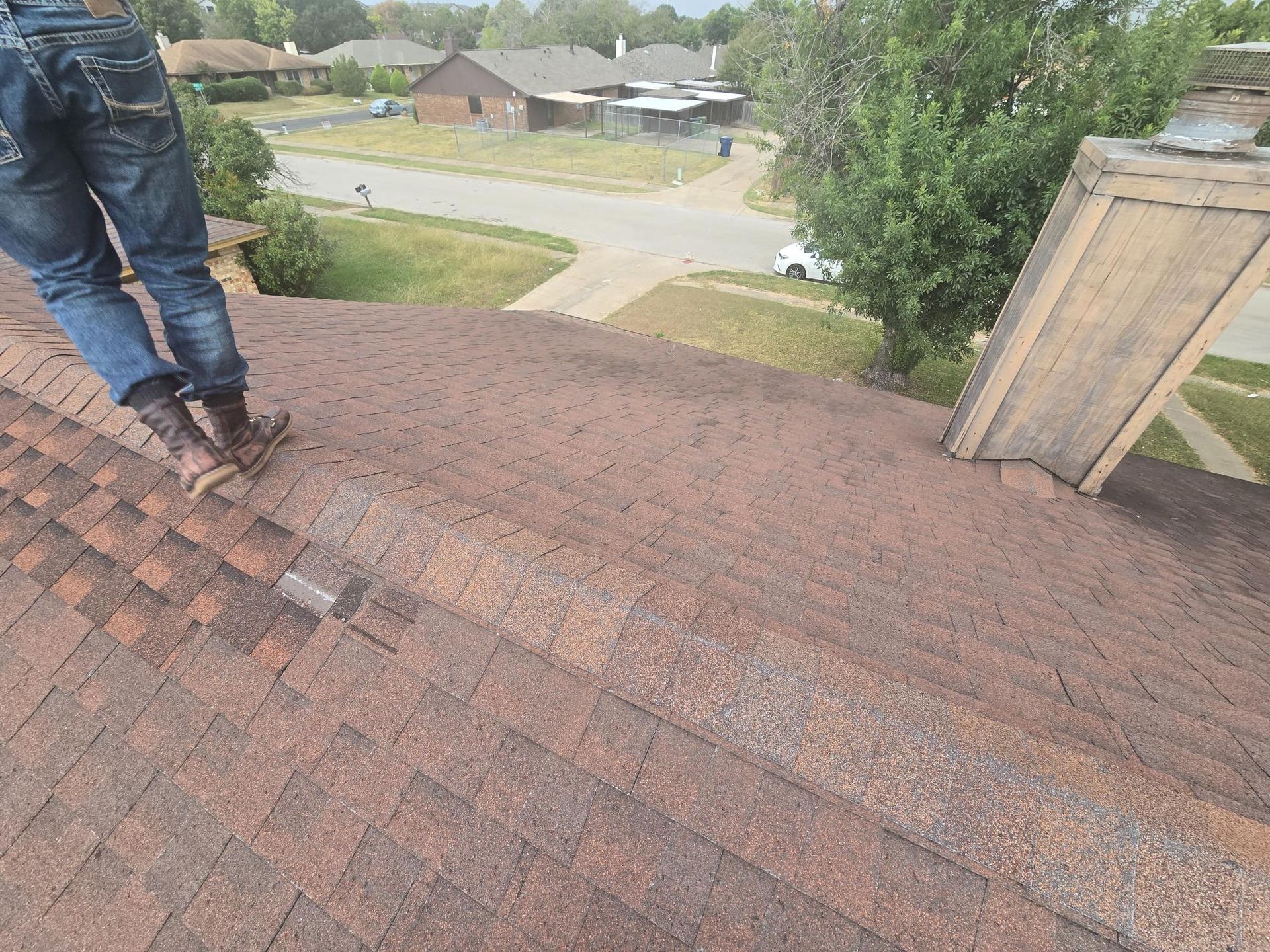 Person on a brown shingle roof inspecting or repairing it, residential setting.