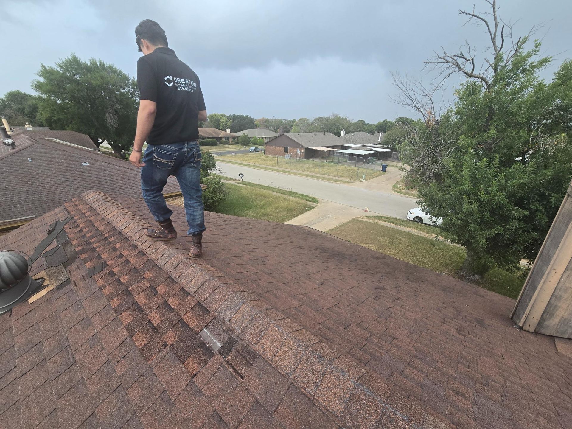 A person in jeans and a black shirt walks on a brown shingled roof, inspecting it. Overcast sky.