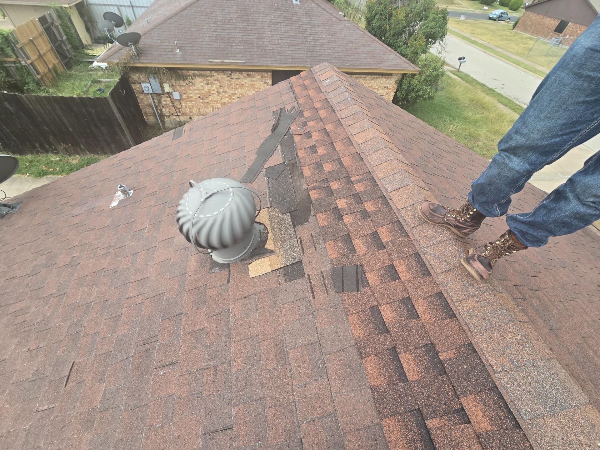 Person standing on a roof with damaged shingles near a vent fan.