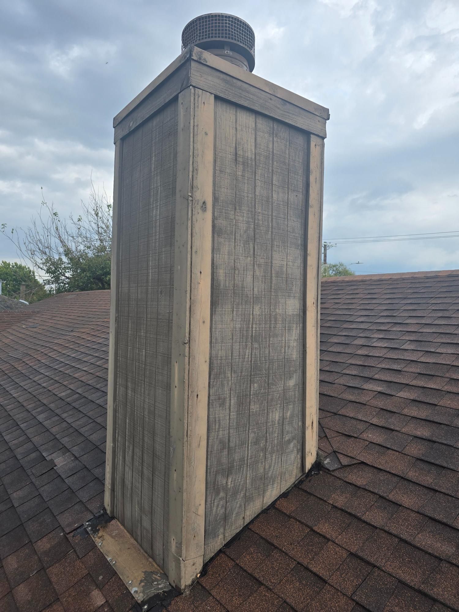 Gray wooden chimney on a brown shingled roof, topped with a metal cap.