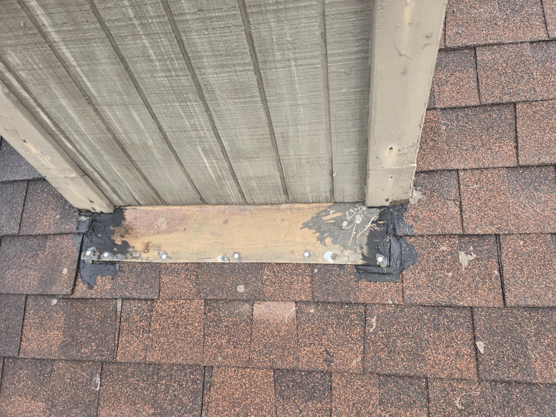 Close-up of a roof with weathered shingles, a wood trim piece, and black sealant where the two meet.