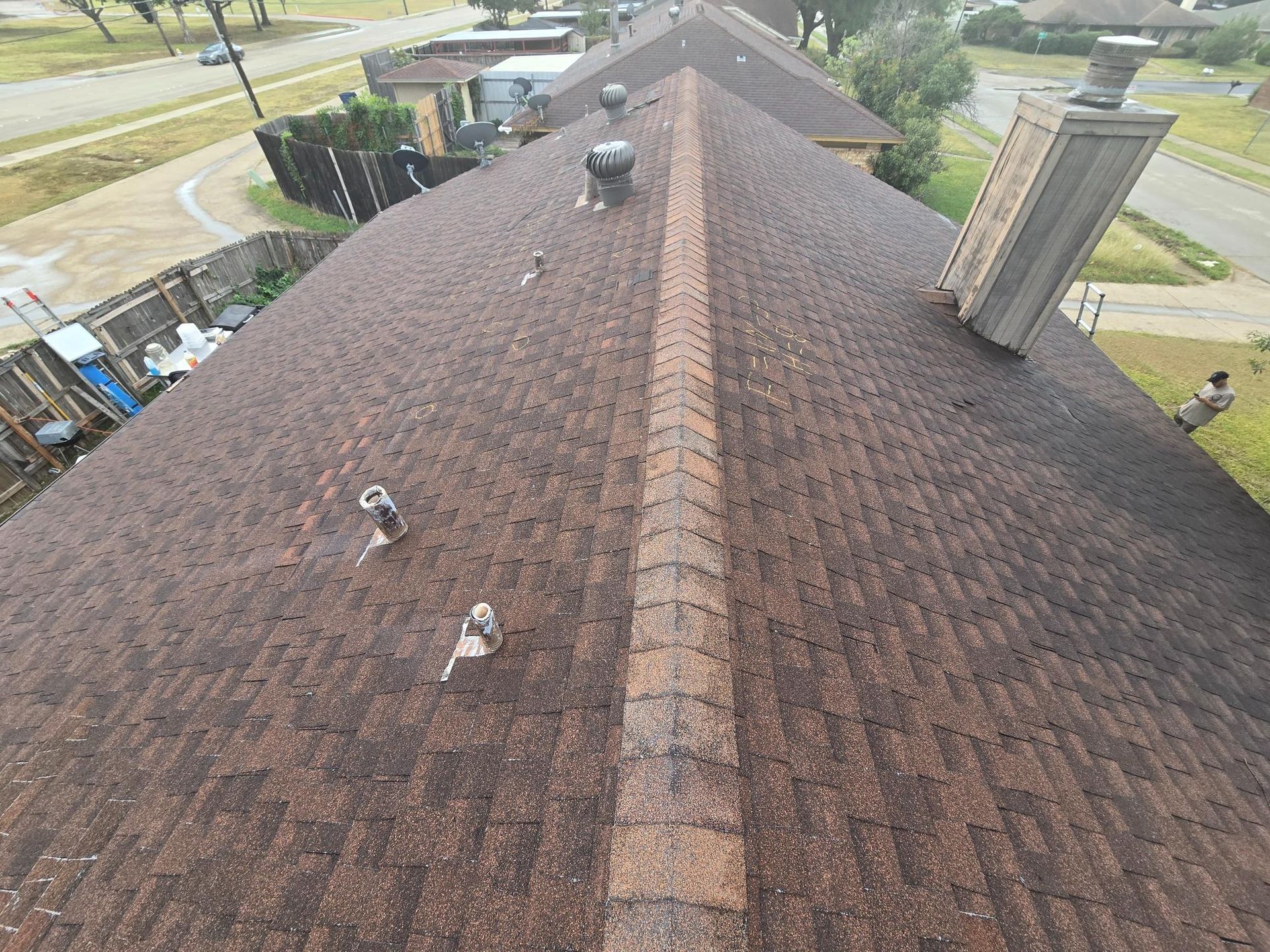 View from above of a brown shingled roof with a brick ridge, various vents, and a chimney.