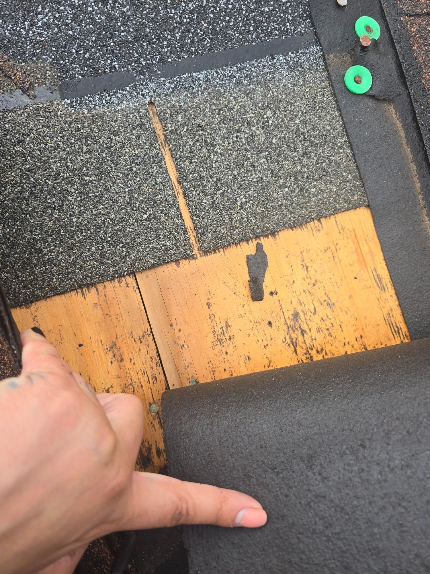 A hand points to a damaged asphalt shingle on a roof, revealing yellow underlayment.