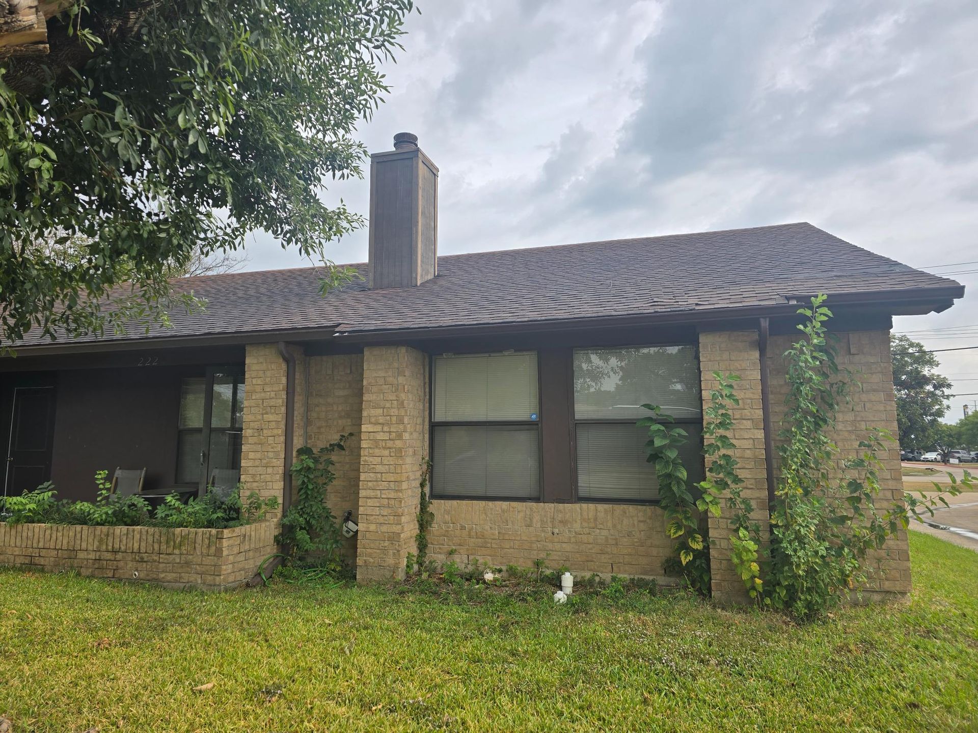 Brick house with dark roof and chimney, overgrown with some greenery; cloudy sky.