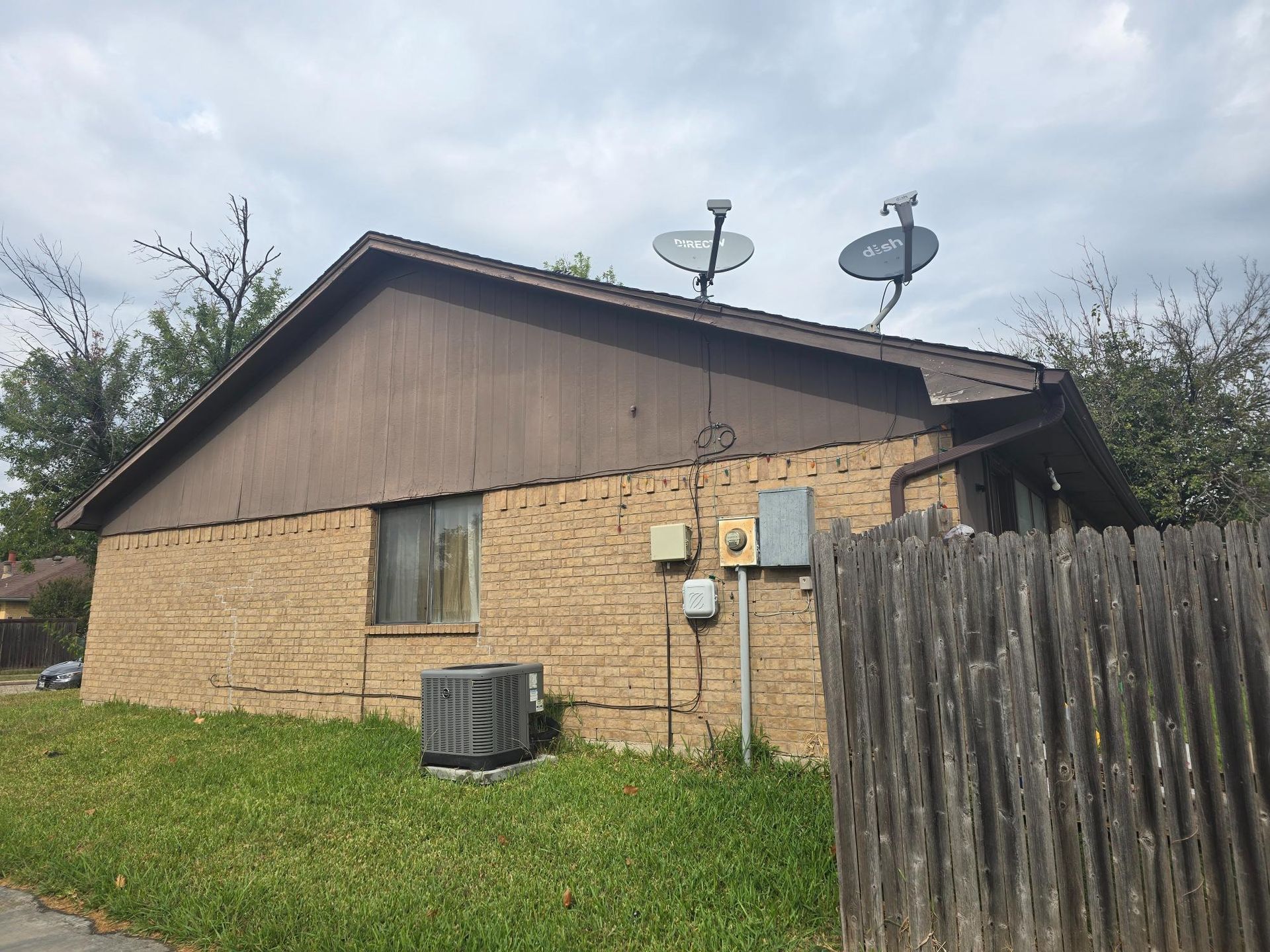 Brick building with brown roof, satellite dishes, air conditioner, and wooden fence.